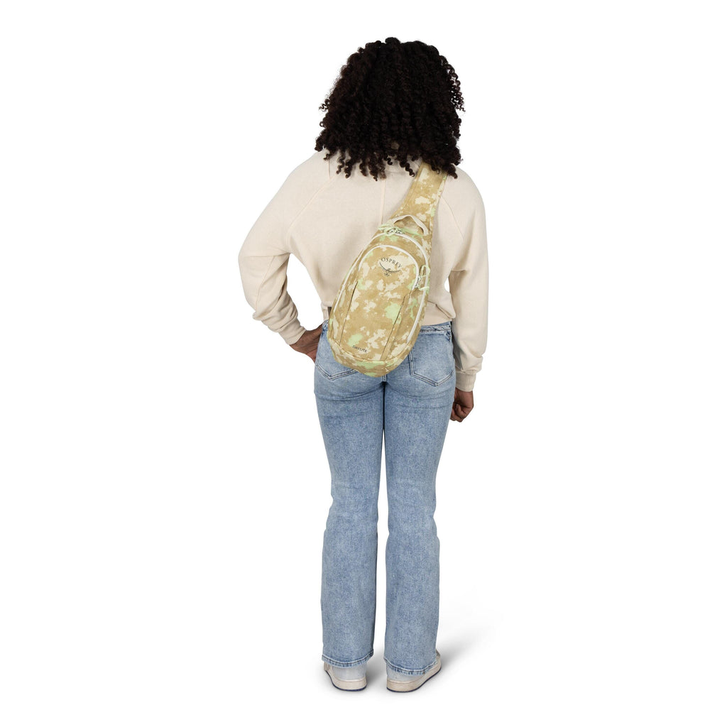 A person is shown from behind, standing with one hand on their hip. They wear a cream sweatshirt, light blue flared jeans, and white sneakers. A tan and mint-green floral sling bag with a diagonal strap is worn across their back. The background is plain w