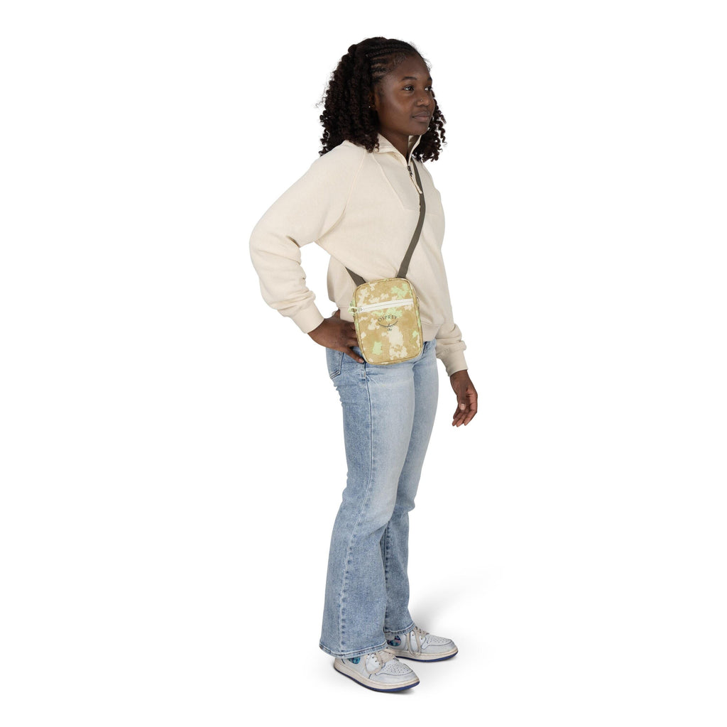 A person stands in a casual pose wearing a cream quarter_zip sweatshirt, light blue flare jeans, and white sneakers with blue accents. They carry a small crossbody bag with a beige and light green floral pattern and olive strap, resting at the hip. One ha