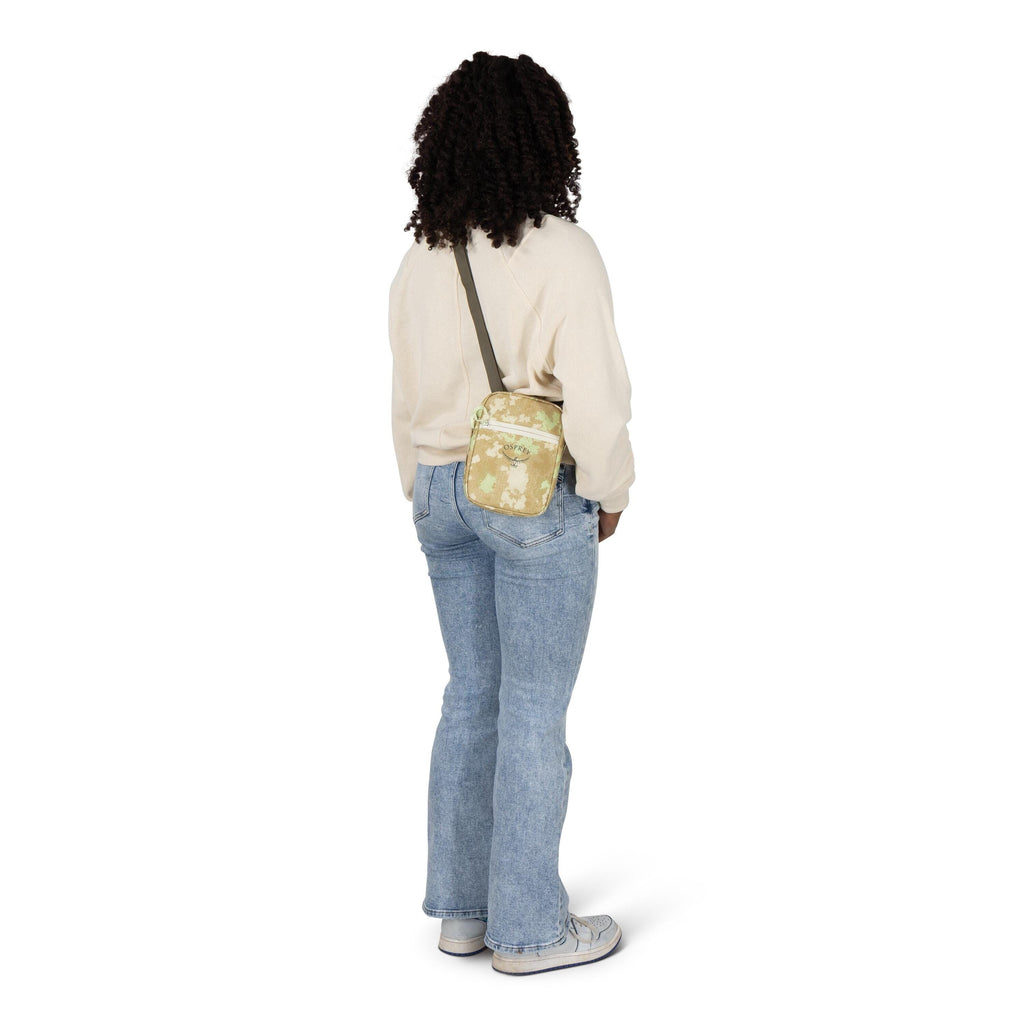 A person is standing with their back slightly turned, wearing a cream sweatshirt, light blue jeans, and gray sneakers. They have a small crossbody bag in a tan camouflage pattern with a dark strap resting on their hip. The background is plain white, keepi