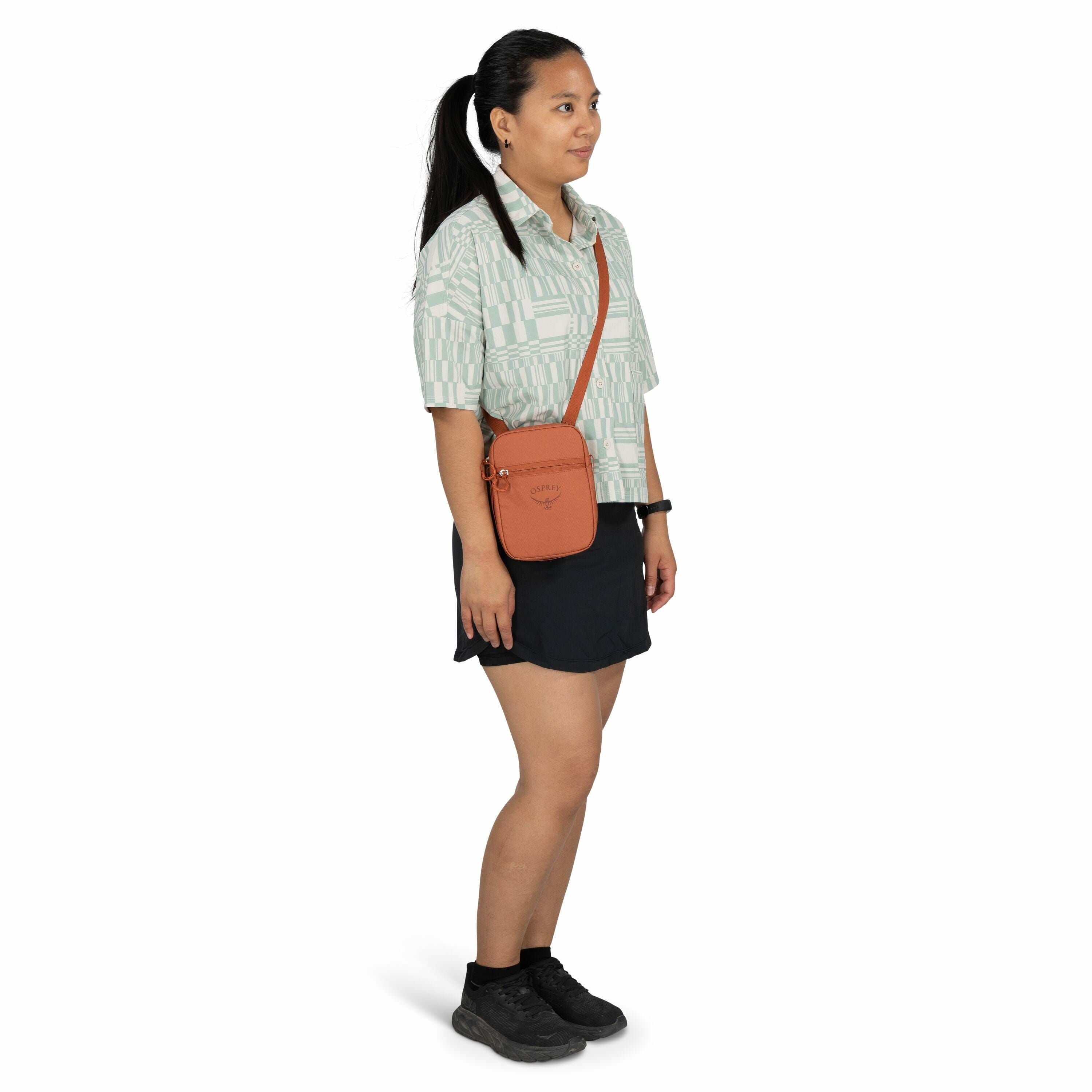 The image depicts a person standing with a casual and sporty outfit. They are wearing a light green patterned shirt with short sleeves and black shorts. The individual has a small orange crossbody bag with the Osprey logo prominently visible, adding a pop