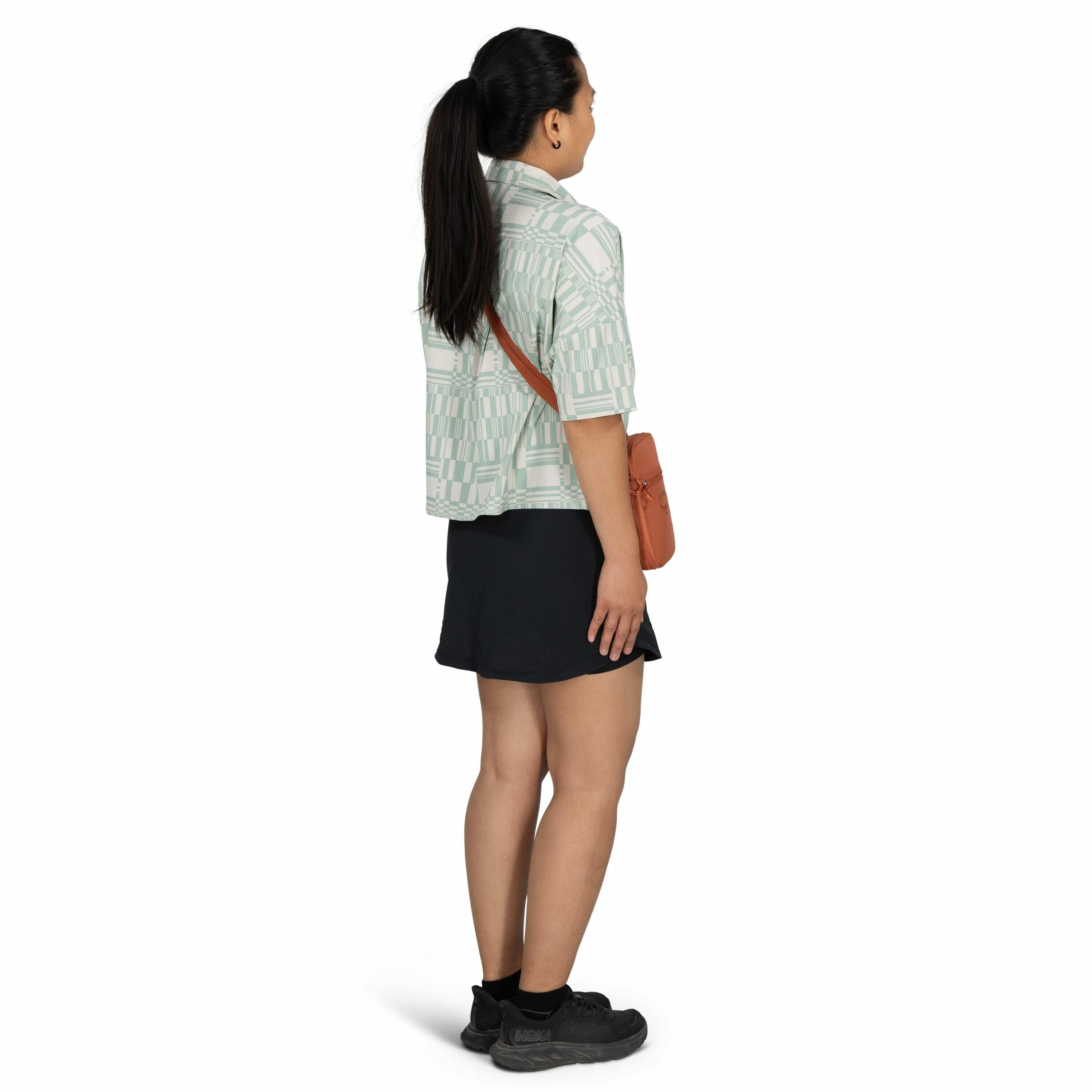 The image shows a person standing with their back facing the camera. They have long, dark hair tied in a ponytail and are wearing a loose-fitting, patterned shirt in light green tones. The person has a brown crossbody bag slung across their body and is dr