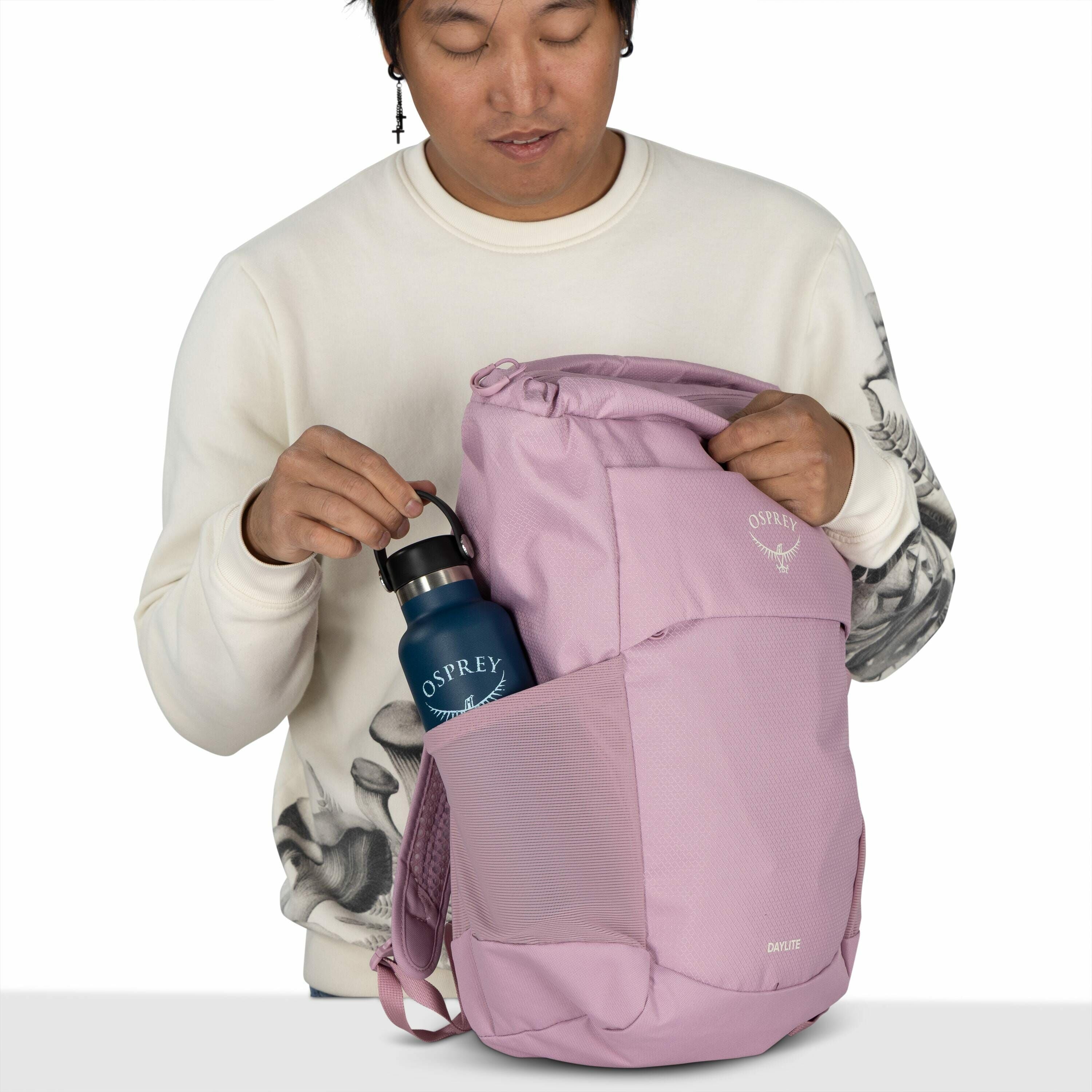 The image shows a person holding a light purple Osprey backpack in one hand while the other hand is placing a blue Osprey water bottle into the mesh side pocket of the backpack. The backpack has the Osprey logo and the word "Daylite" printed on it, indica