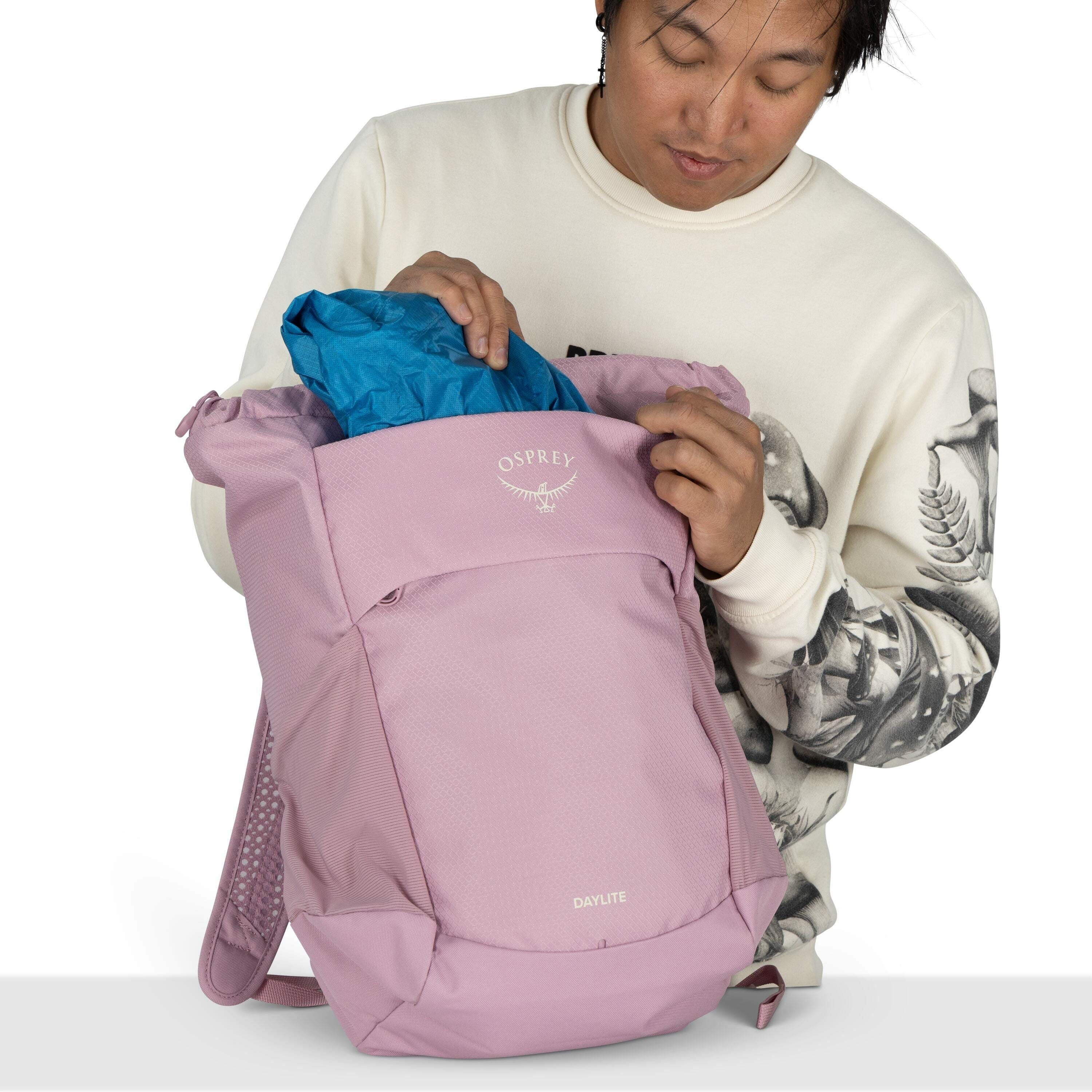 The image shows a person holding a backpack made by the brand Osprey. The backpack is light pink and has "OSPREY" and the logo of an osprey bird printed in white on the front. The backpack model name "DAYLITE" is also visible near the bottom. The person i
