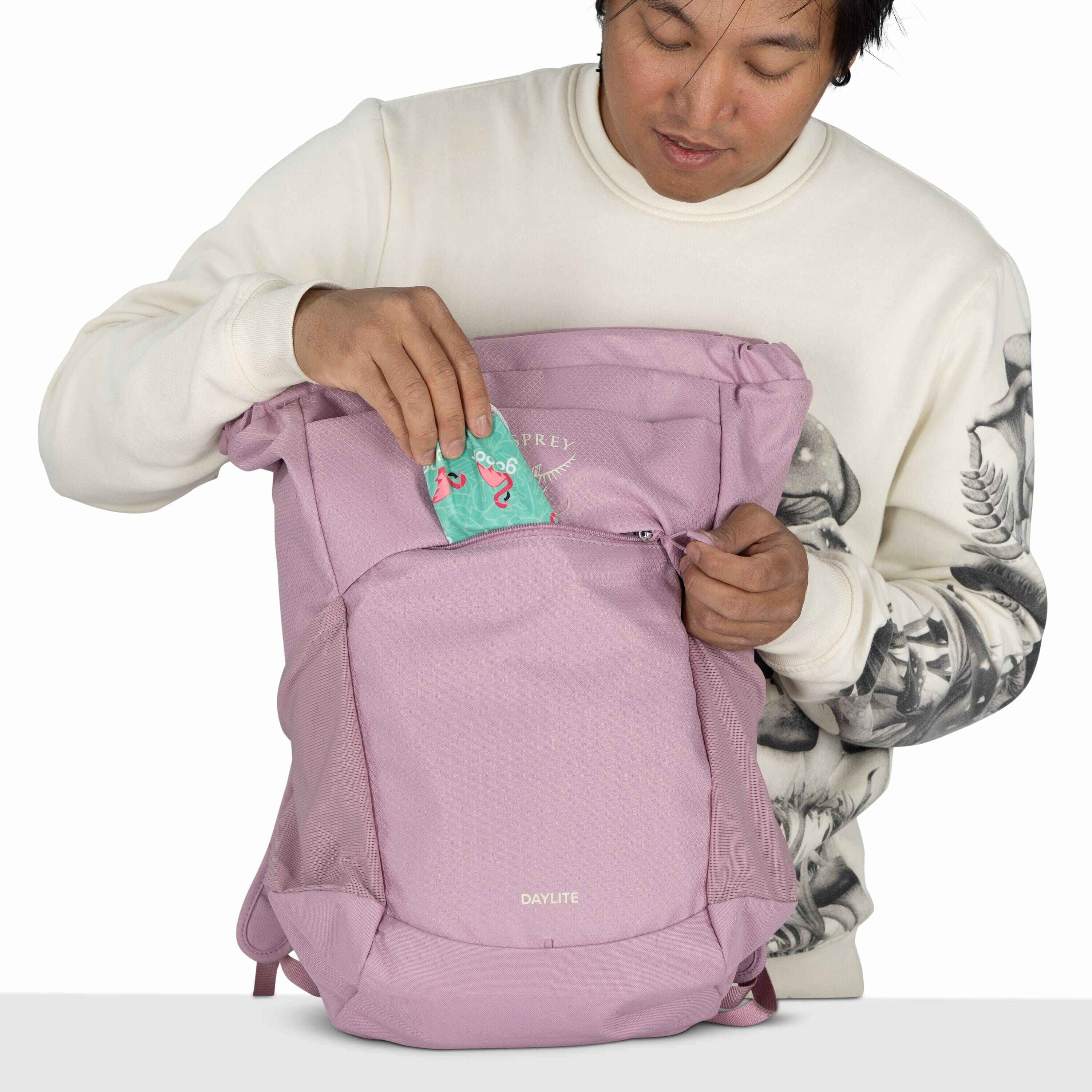 The image features a person holding a light purple Osprey Daylite backpack. The individual is placing a colorful package, which features a cartoon-style illustration of flamingos on a turquoise background, into the front pocket of the backpack. The person
