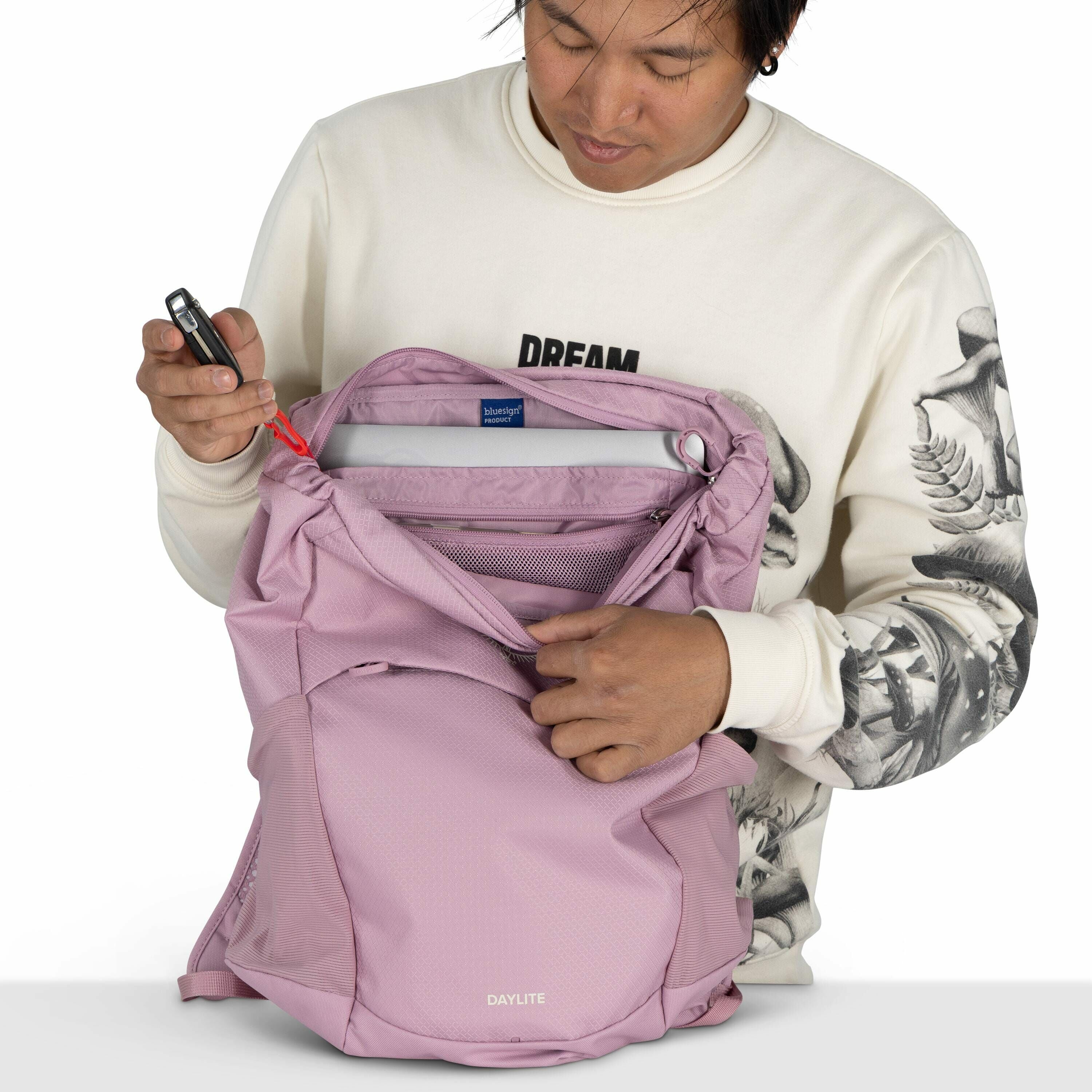 The image shows a person wearing a white sweatshirt with black and white floral patterns on the sleeves, and the word "DREAM" visible on the chest. They are holding open a pink Daylite backpack, revealing its interior compartments. Inside the backpack, th