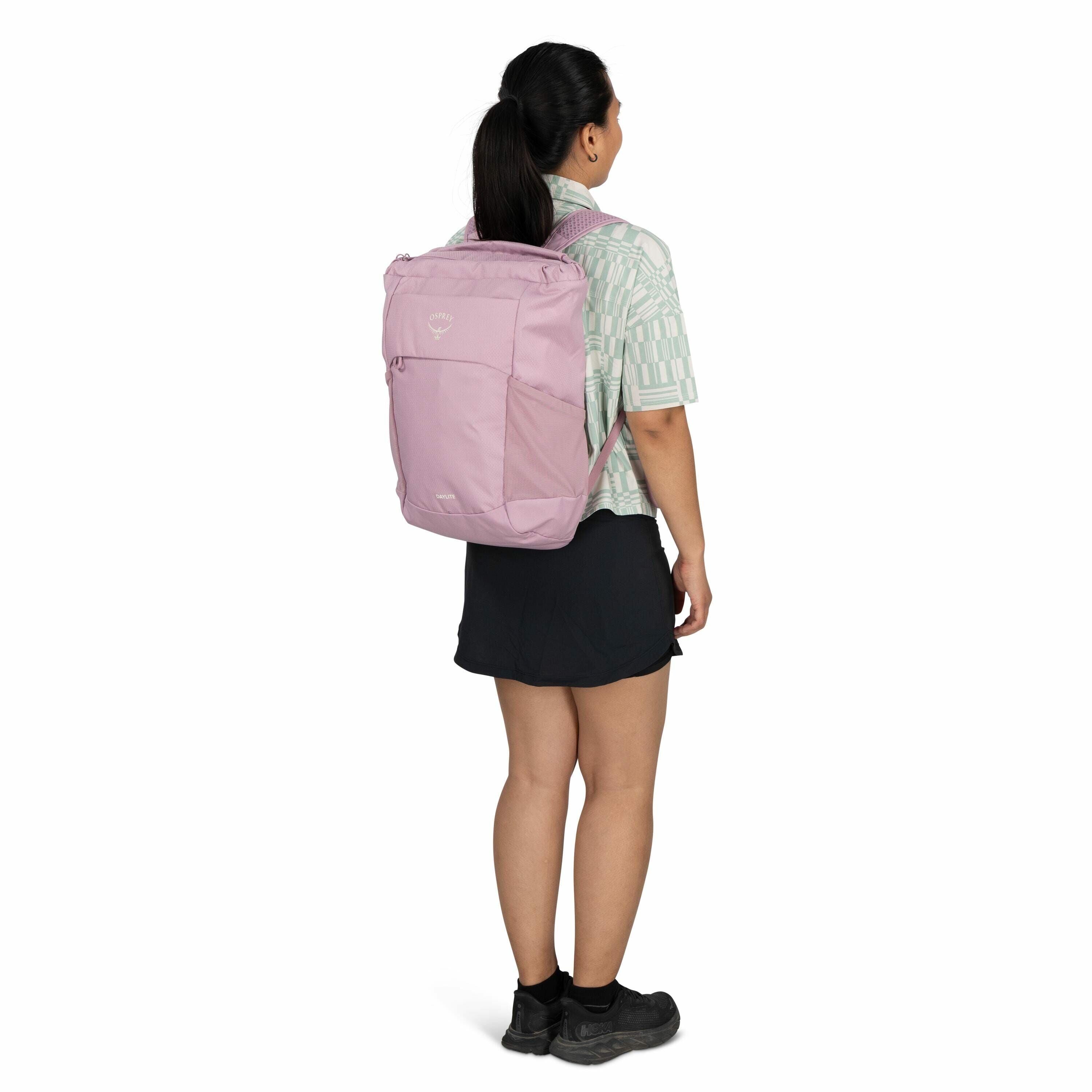 The image shows a woman standing and facing slightly to the left, wearing a light pink Osprey backpack on her back. The backpack has a visible Osprey logo and the model name "Arcane" printed in white. She is dressed in a light green patterned short-sleeve