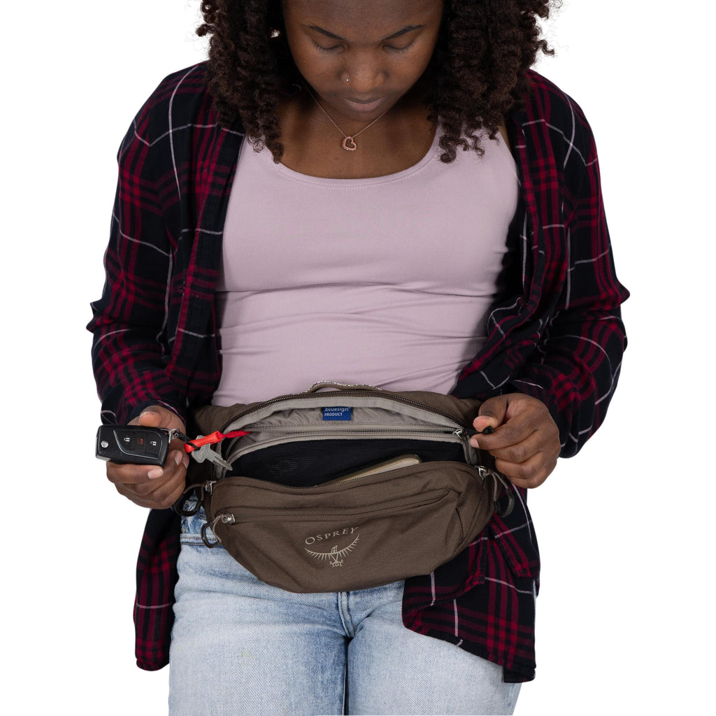 A person wearing a plaid shirt over a mauve tank top and light jeans is shown from the shoulders down. They are holding open a brown Osprey waist pack with the main zipper unzipped, revealing items inside including a phone and other essentials. A red key
