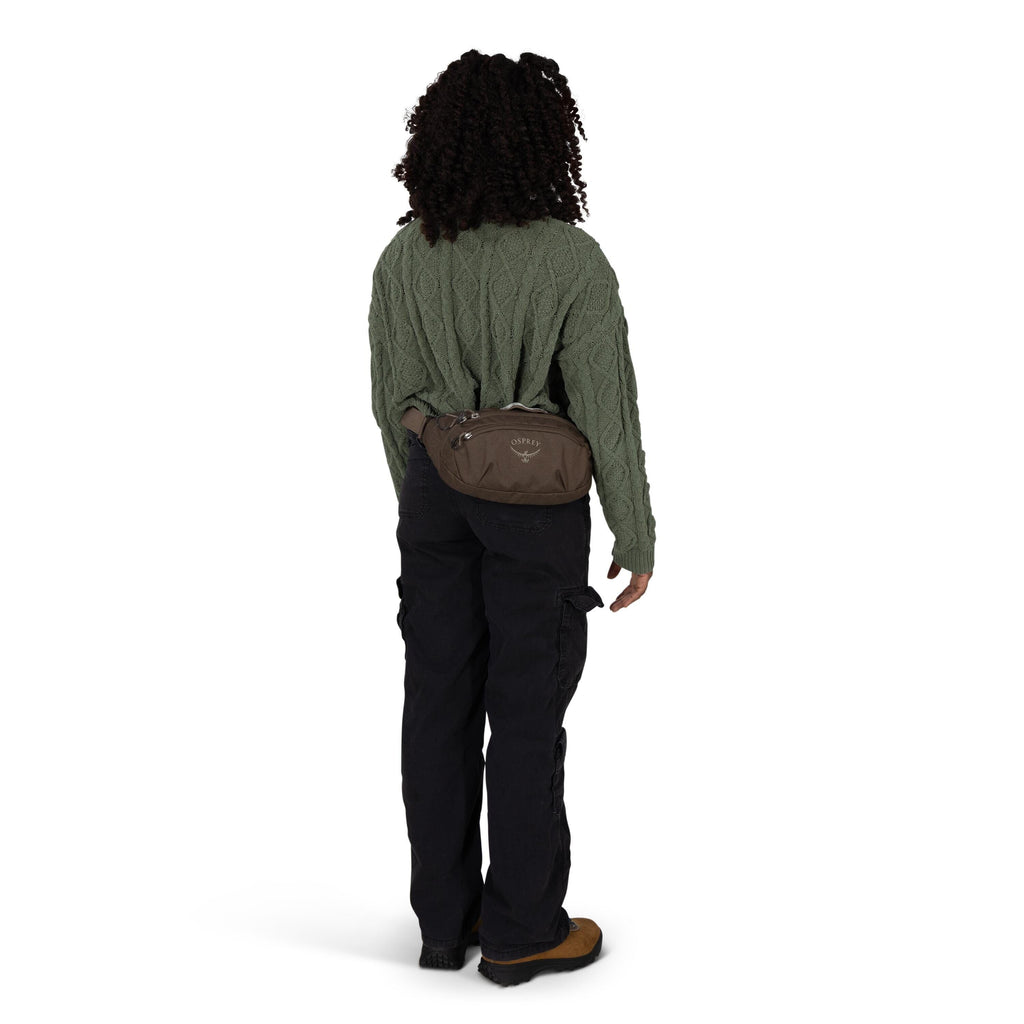 A person is shown from the back wearing a textured, cable-knit green sweater, black cargo pants, and brown hiking shoes. They have a brown Osprey waist pack strapped around their hips, with the logo visible on the pouch. The individual has curly hair and