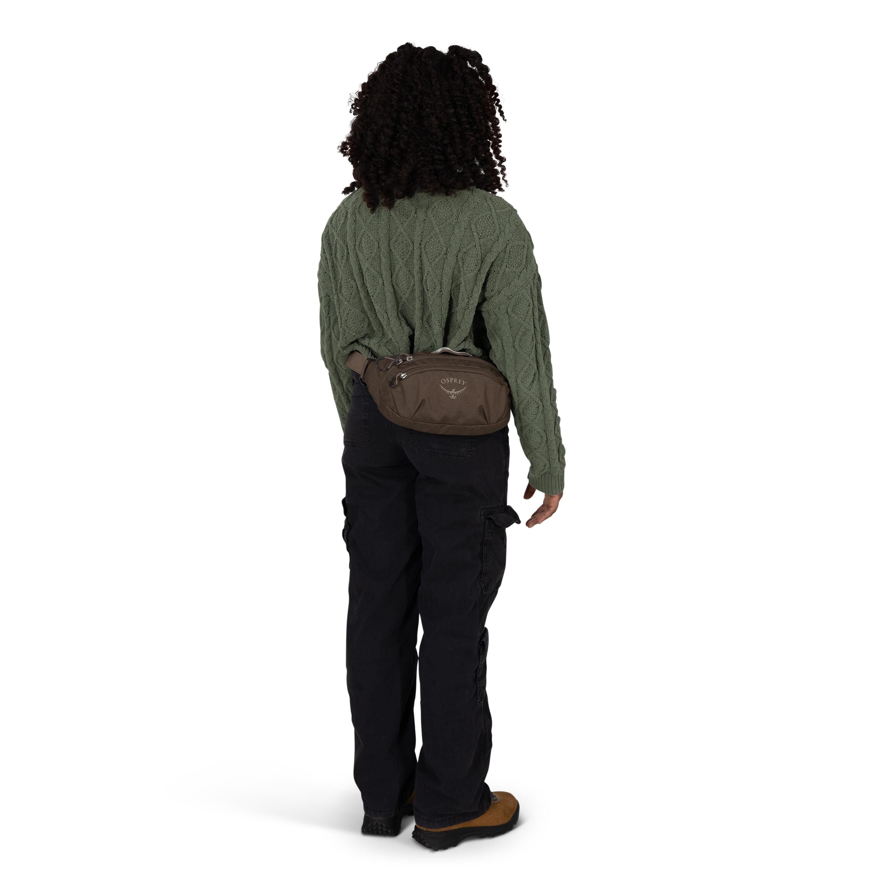 A person is shown from the back wearing a textured, cable-knit green sweater, black cargo pants, and brown hiking shoes. They have a brown Osprey waist pack strapped around their hips, with the logo visible on the pouch. The individual has curly hair and
