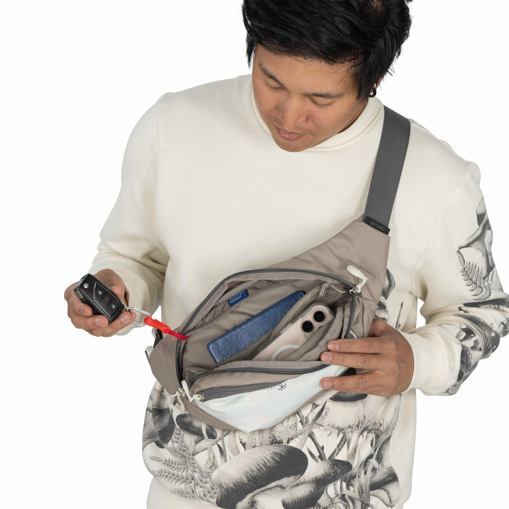 The image depicts a person wearing a light-colored sweater with a black and white mushroom print on the sleeves and lower section. They have a gray crossbody bag worn across their chest, which is unzipped to reveal its contents. Inside the bag, there's a