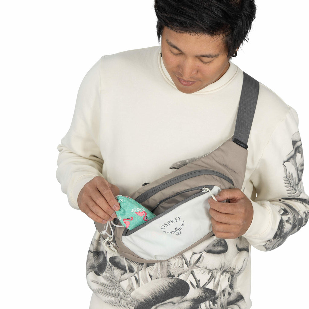 The image shows a person wearing a cream-colored sweatshirt with detailed mushroom illustrations. They are also wearing a gray Osprey sling bag across their body. The person is in the process of placing or removing a colorful item, possibly a can or a sna