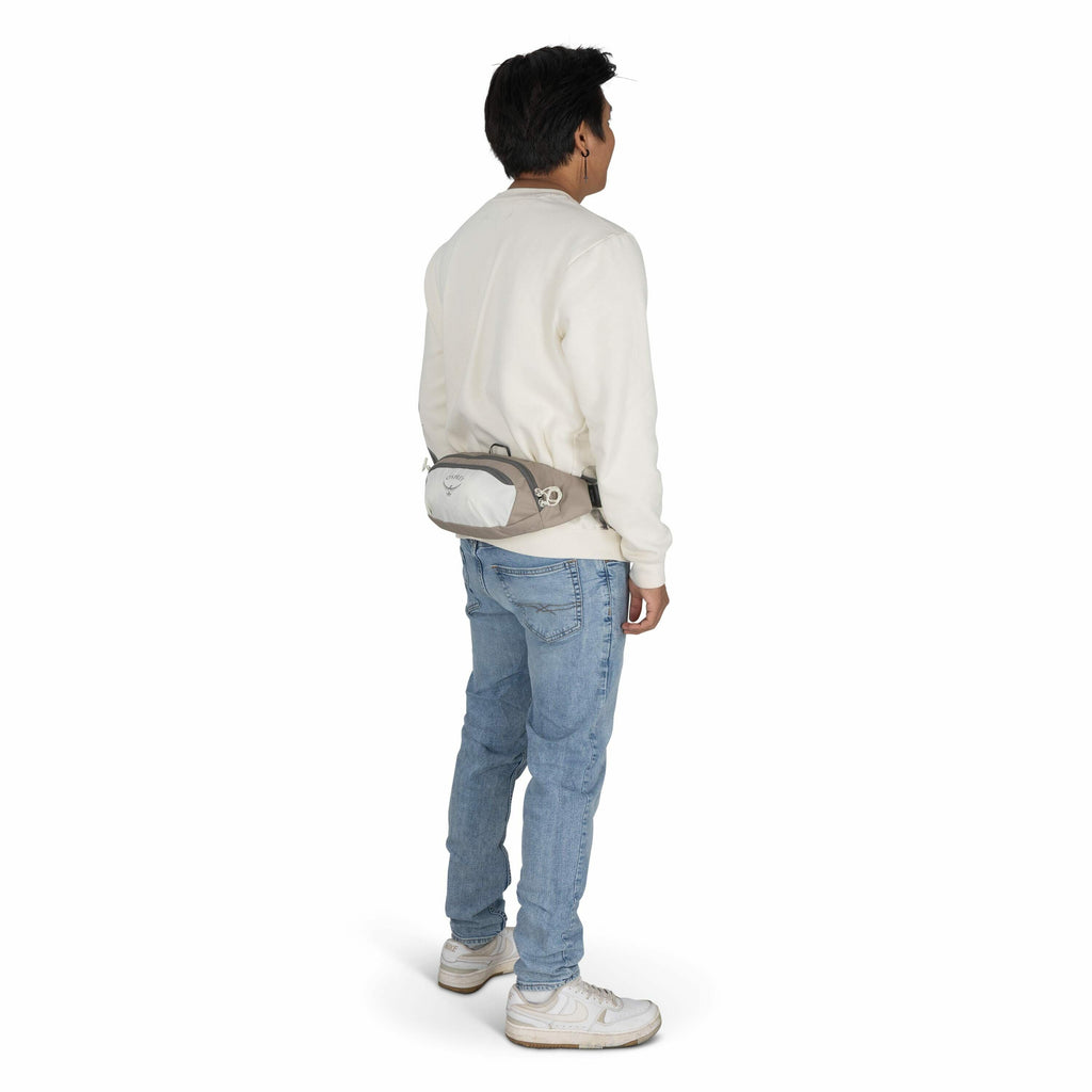The image features a man standing facing away from the camera. He is wearing a cream-colored sweatshirt, light blue jeans, and white sneakers. Around his waist is a small gray and white pouch with the Osprey logo, which resembles a stylized bird emblem, s