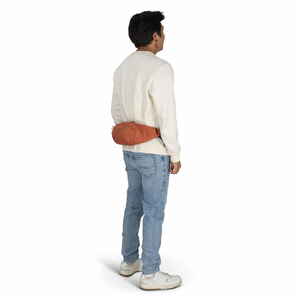 The image shows a young man standing in a relaxed pose, facing slightly to the right, with his back visible. He is wearing a cream-colored long-sleeve shirt, light blue jeans, and white sneakers. Around his waist, he has a rust-colored Arc'teryx hip pack,