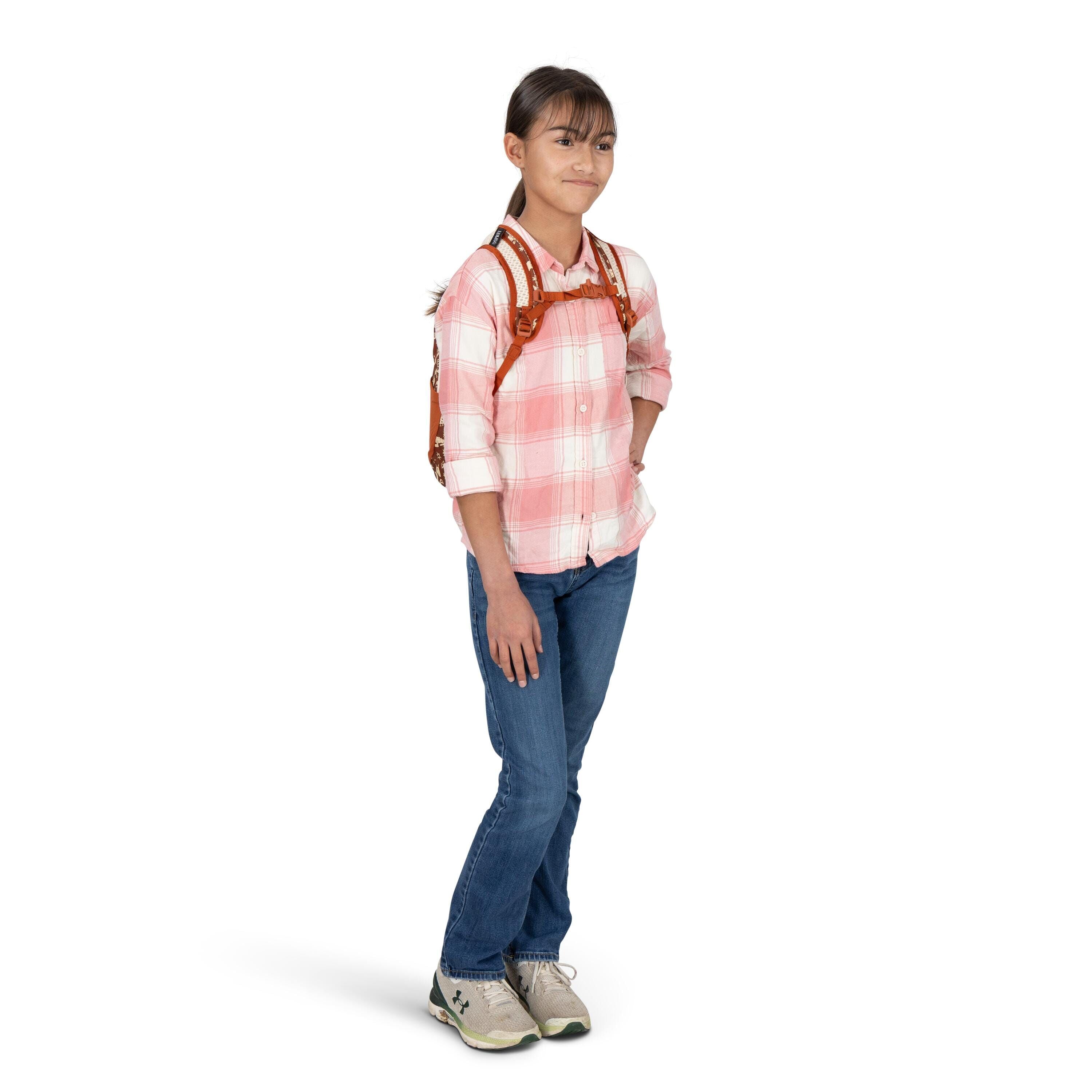 A person stands casually with one hand in their pocket, wearing a pink-and-white plaid button-up shirt and blue jeans. They have a brown leather-style backpack with visible decorative studs and straps. The person is wearing light-colored athletic shoes wi