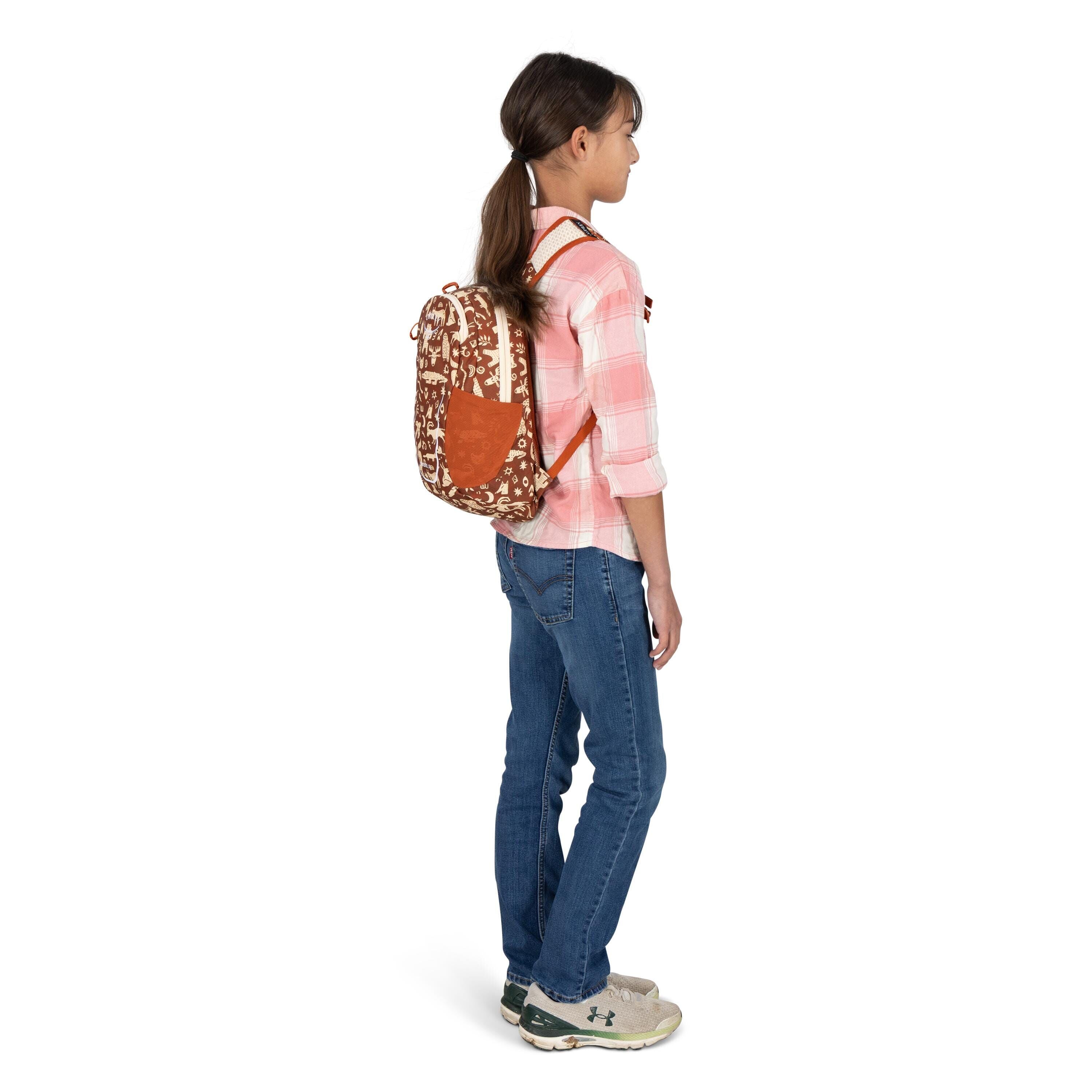 A young person stands in profile wearing a small backpack with a brown and white floral pattern and orange accents. They have a pink-and-white plaid shirt with rolled sleeves, blue jeans, and beige Under Armour sneakers. Their hair is tied back in a low p
