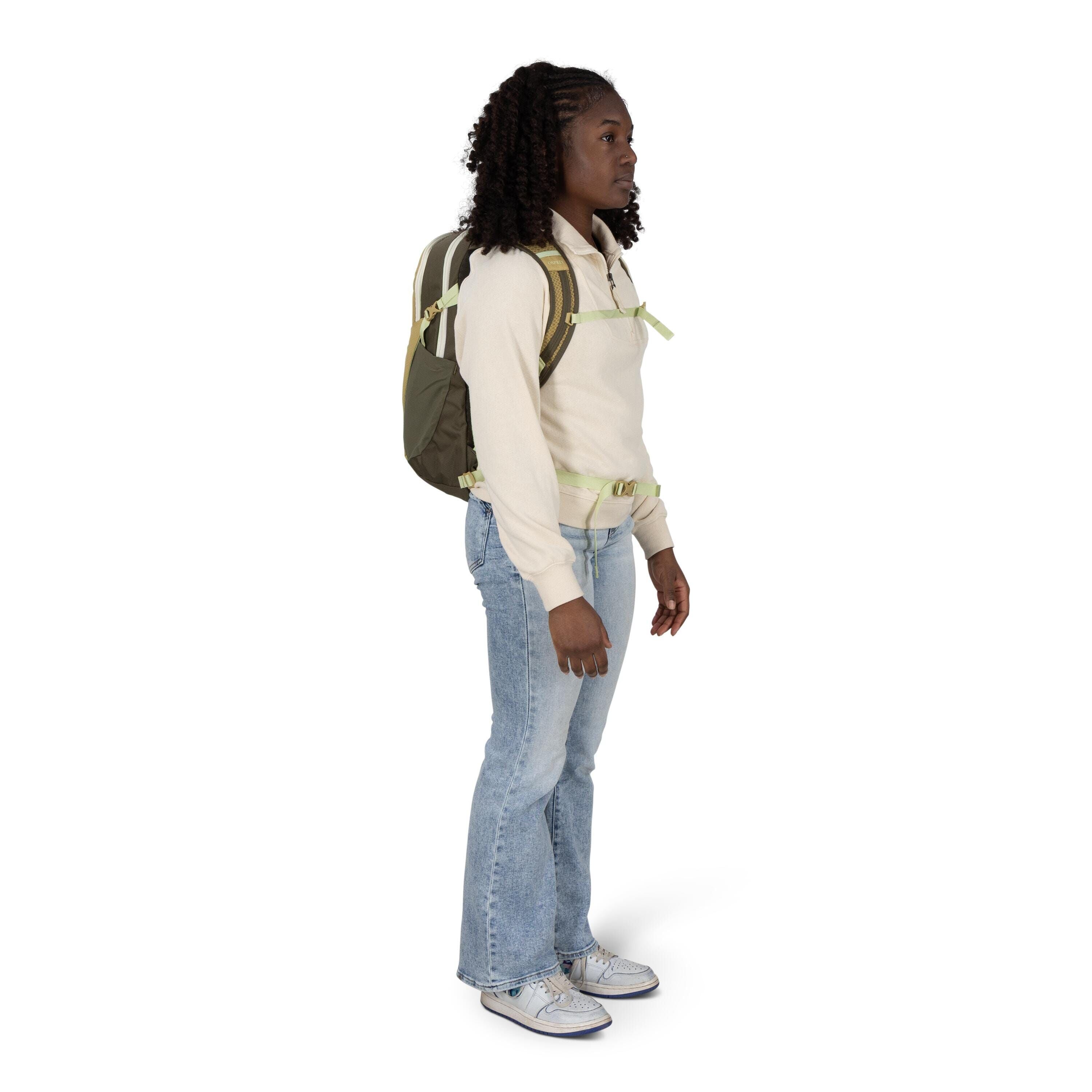 A person stands in profile wearing a light beige pullover, light-wash bootcut jeans, and white sneakers. They have a green backpack with light green straps secured across the chest and waist. Their hair is styled in tight curls, and their hands rest at th