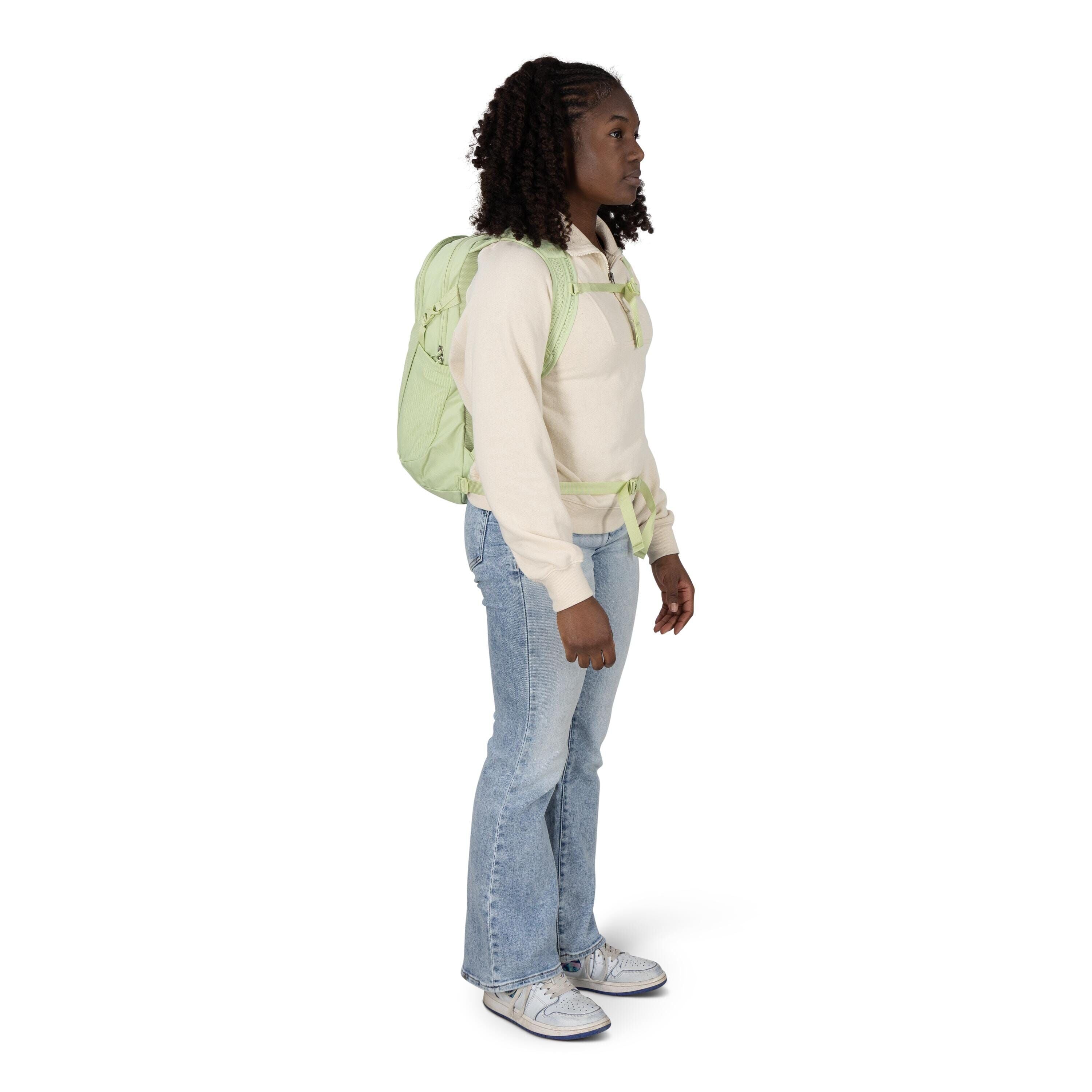 A person stands in profile wearing a casual outfit and a light green backpack. They have a cream-colored pullover with a half-zip, light blue bootcut jeans, and light gray sneakers with blue accents. The backpack's shoulder and chest straps are visible, a