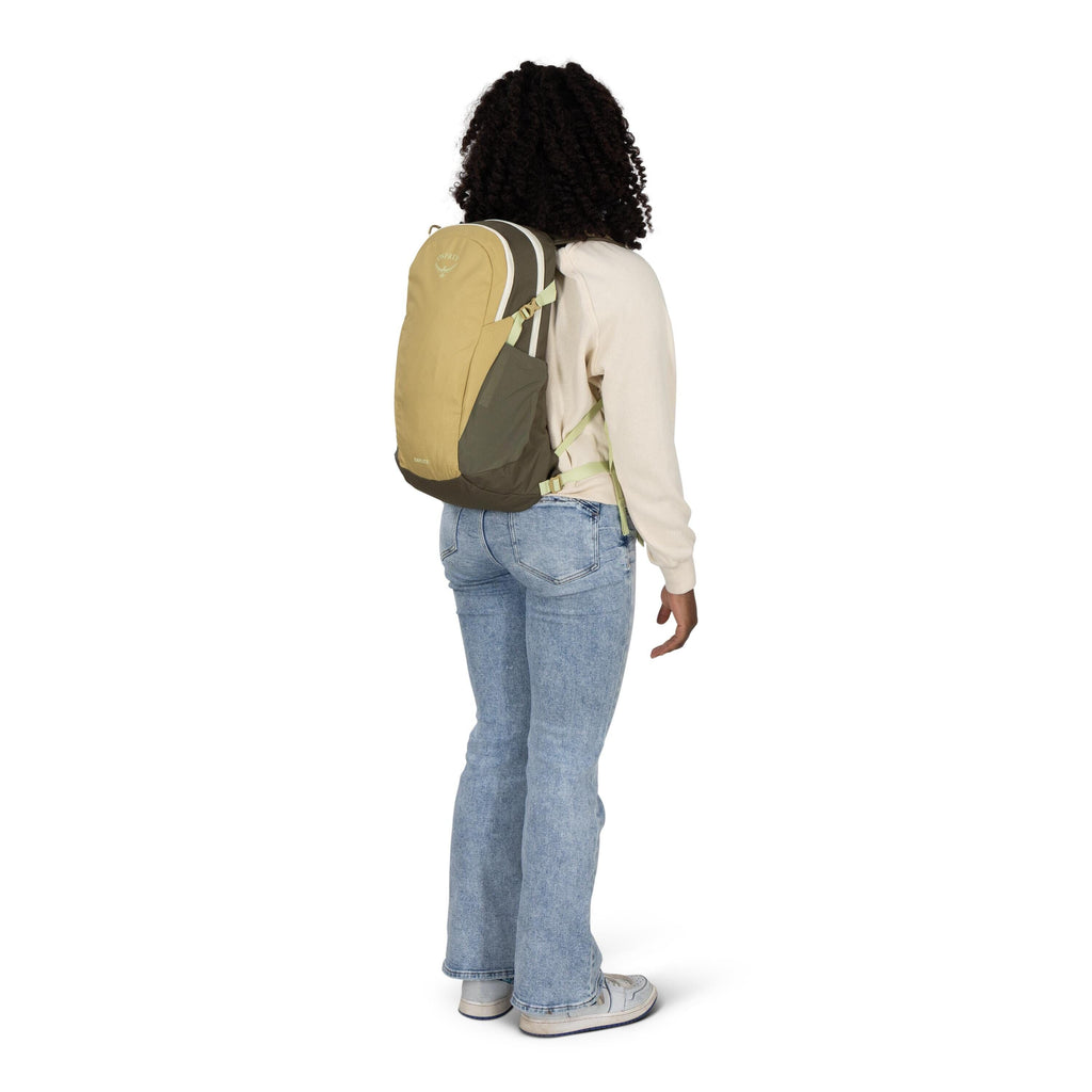 A person is standing turned slightly to the side, wearing a small hiking backpack. The pack is two-tone in tan and olive with side stretch pockets and a light-colored zipper; the Osprey logo is visible near the top. They're dressed in a cream sweatshirt,