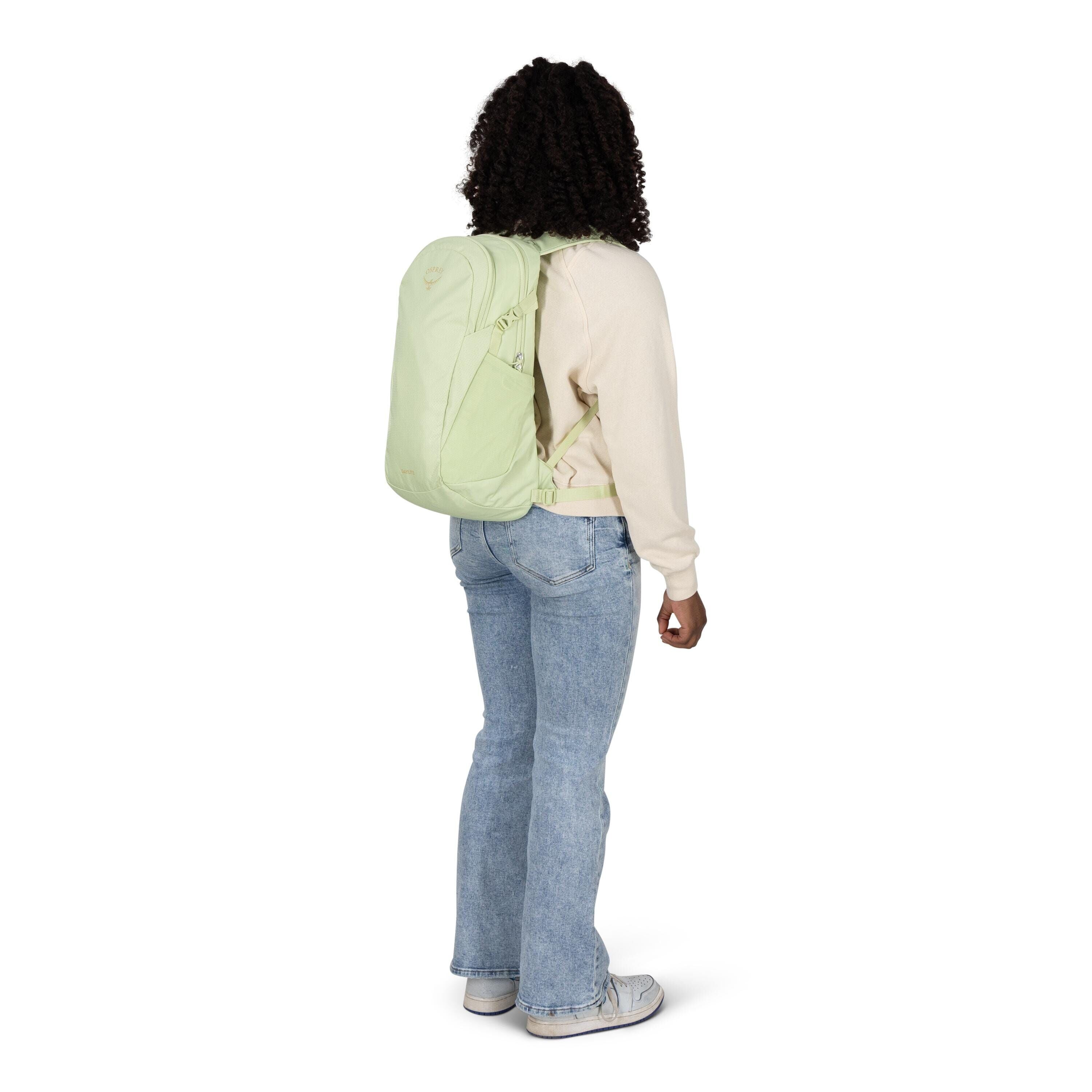 A person is standing and facing slightly away, wearing a light green backpack. They have on a cream long-sleeve top, light blue bootcut jeans, and gray sneakers. The backpack appears streamlined with multiple zippered compartments and side pockets, and it