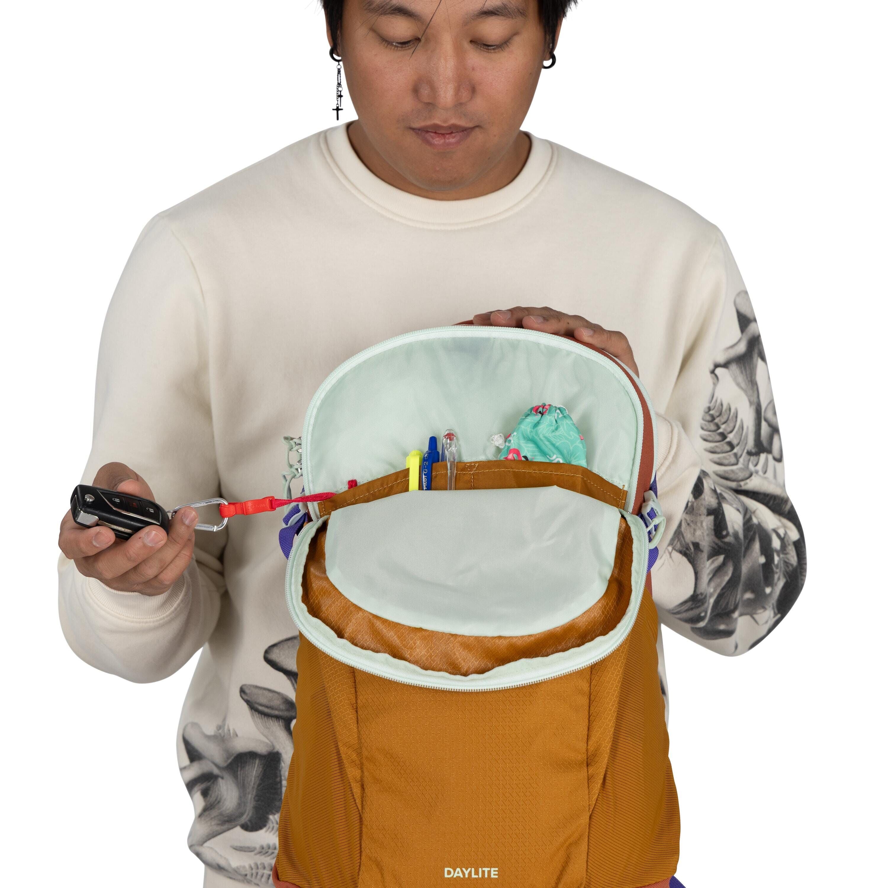 The image depicts a person wearing a cream-colored sweatshirt with intricate black and white designs on the sleeves. The person is holding an orange "DAYLITE" backpack, which is partially open to reveal its contents. Inside the backpack are items like pen