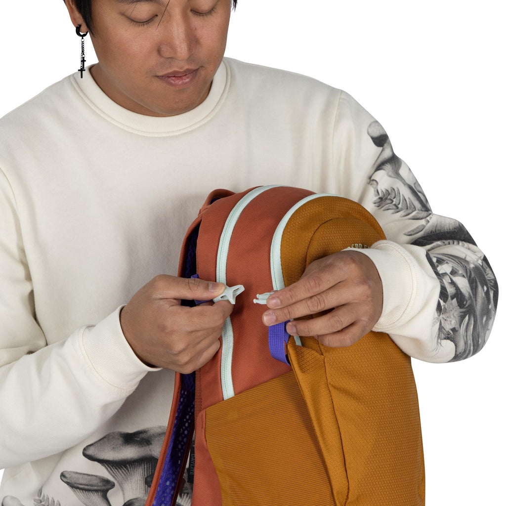 The image shows a person wearing a cream-colored sweatshirt with detailed black-and-white mushroom illustrations on the sleeve. They are holding an orange and brown backpack with mint green zippers and a visible front pocket. The person is in the process