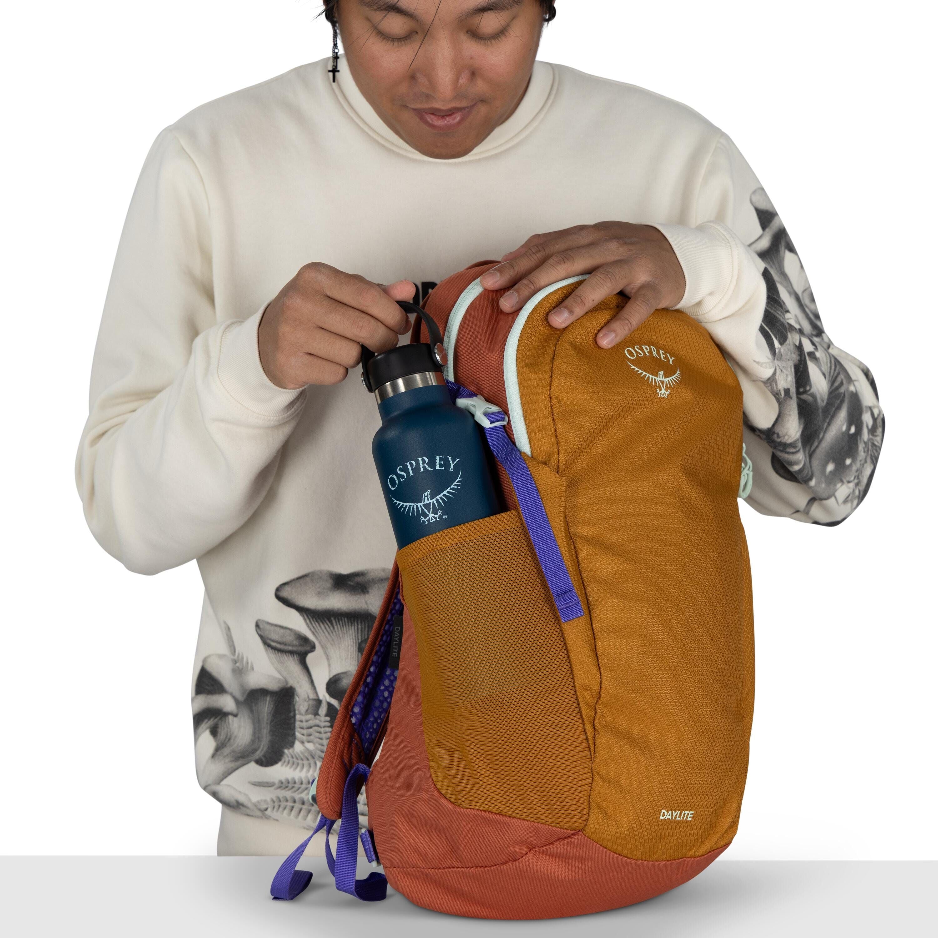The image shows a person wearing a white sweatshirt with a black and white mushroom pattern on the sleeves and chest area. They are holding an Osprey Daylite backpack, which is orange with purple accents. In the side pocket of the backpack, there's a blue