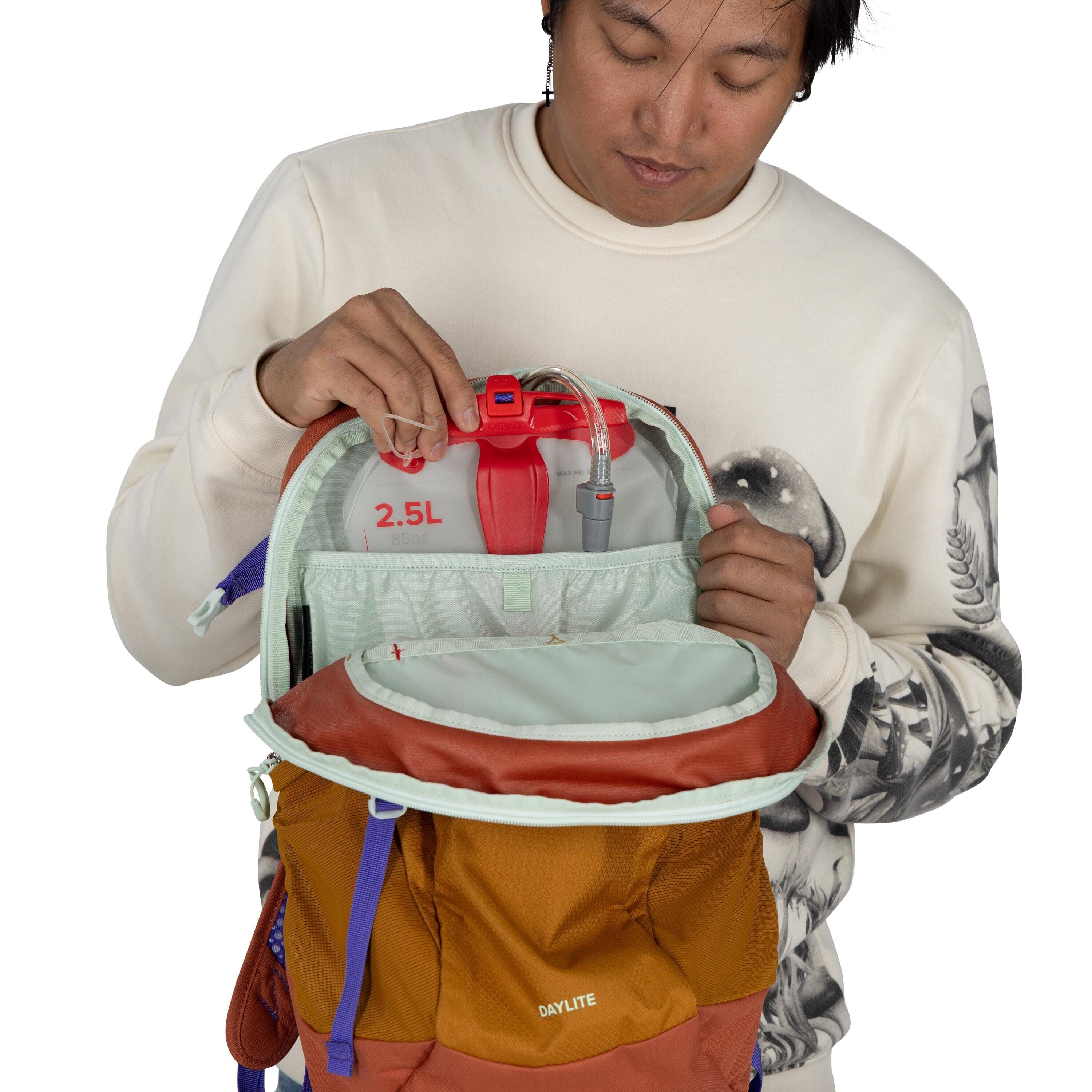 The image shows a person holding open a backpack to reveal its contents. Inside the backpack, there is a hydration reservoir with a capacity of 2.5 liters, indicated by the markings "2.5L 85oz" in red text. The backpack is a multi-colored design, primaril