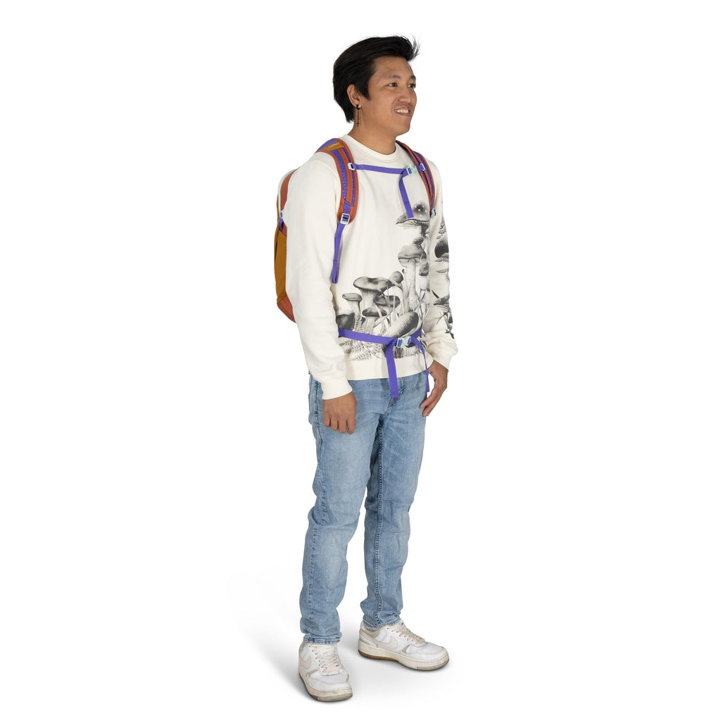 The image shows a young person standing and dressed casually. They are wearing a white sweater with a distinctive mushroom print, paired with light blue jeans and white sneakers. A colorful backpack with orange, yellow, and purple accents is worn over bot