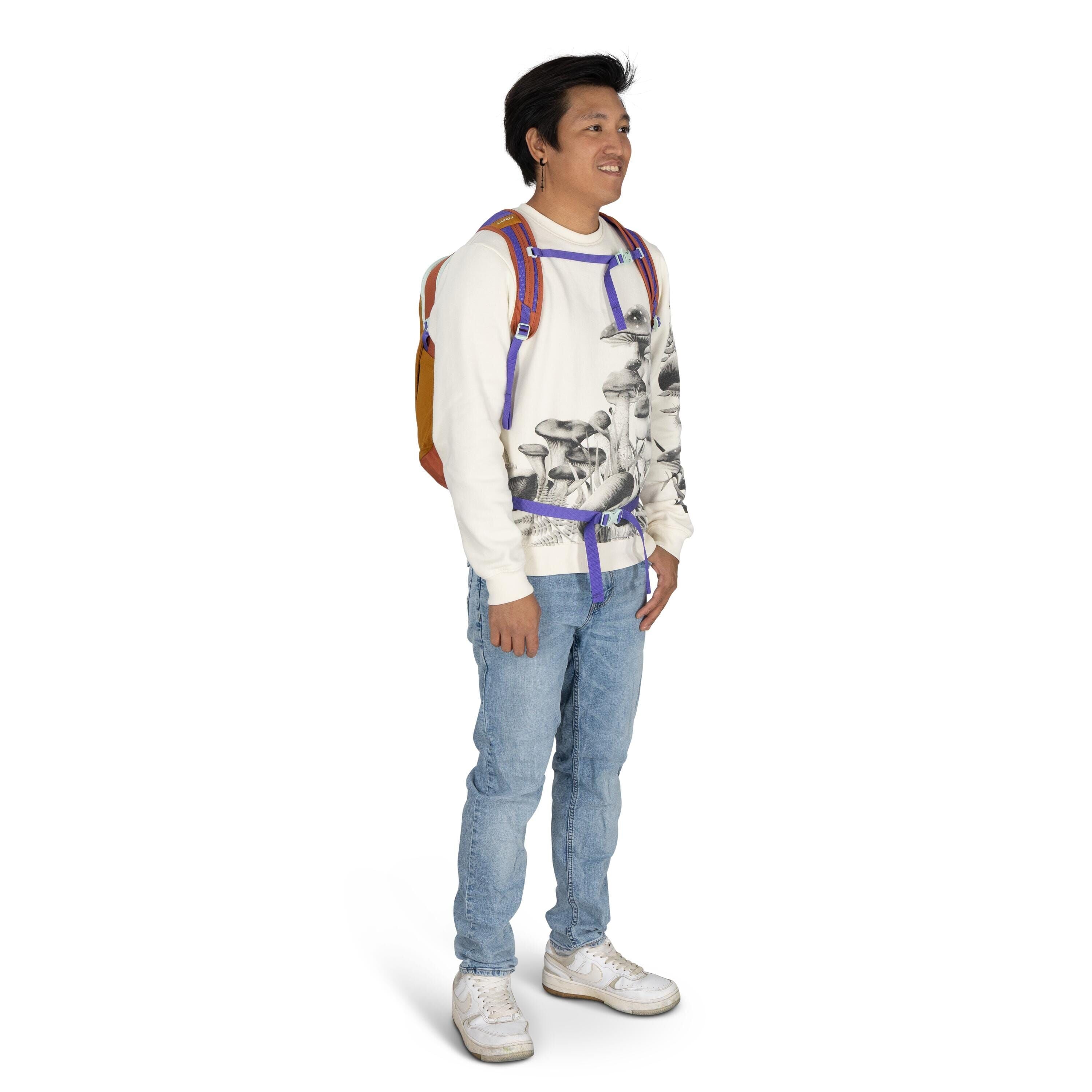 The image shows a young person standing and dressed casually. They are wearing a white sweater with a distinctive mushroom print, paired with light blue jeans and white sneakers. A colorful backpack with orange, yellow, and purple accents is worn over bot