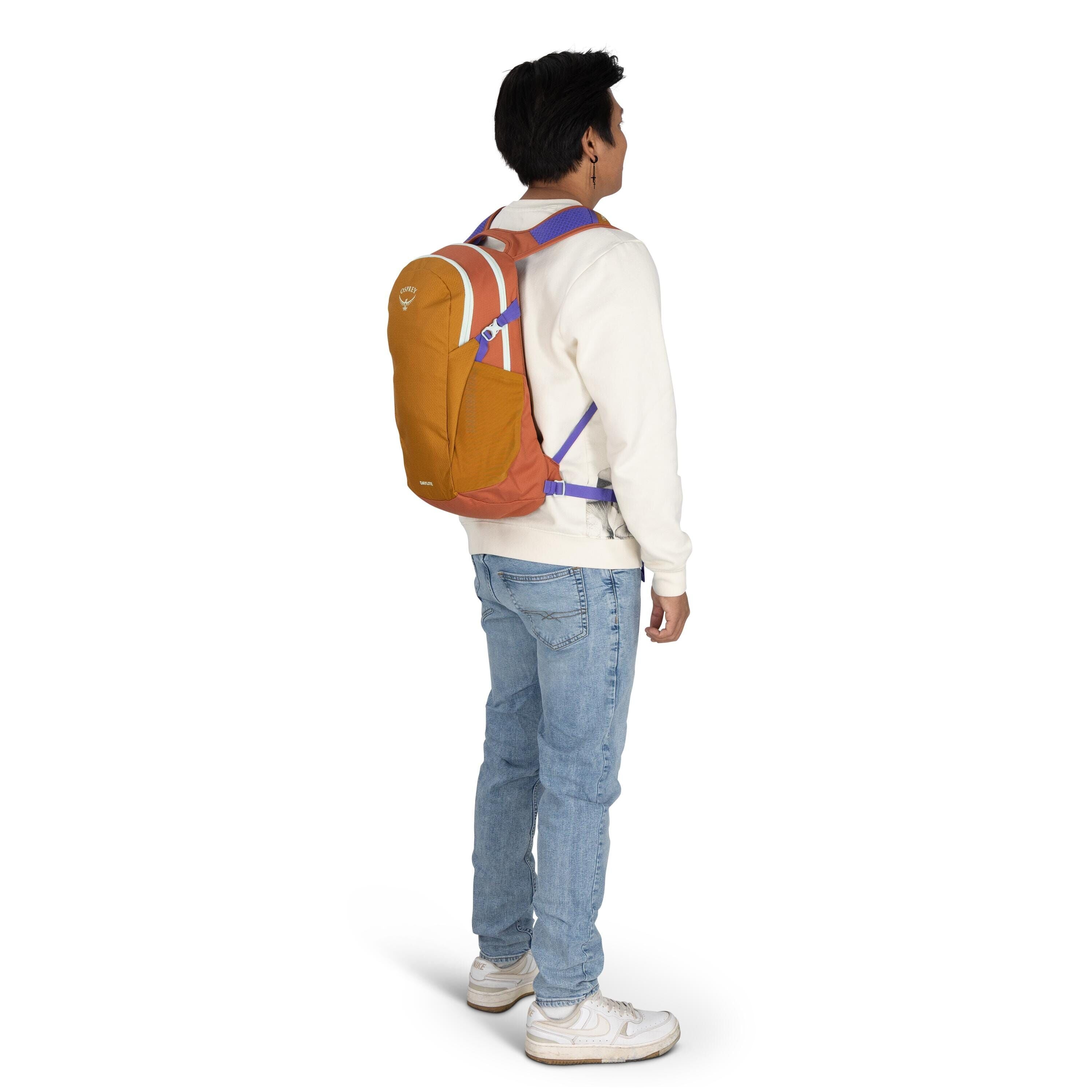 The image shows a young man facing away from the camera, wearing a casual outfit comprised of a white sweater and light blue jeans. He has on white sneakers, which complement the overall casual look. On his back is a colorful backpack with a combination o