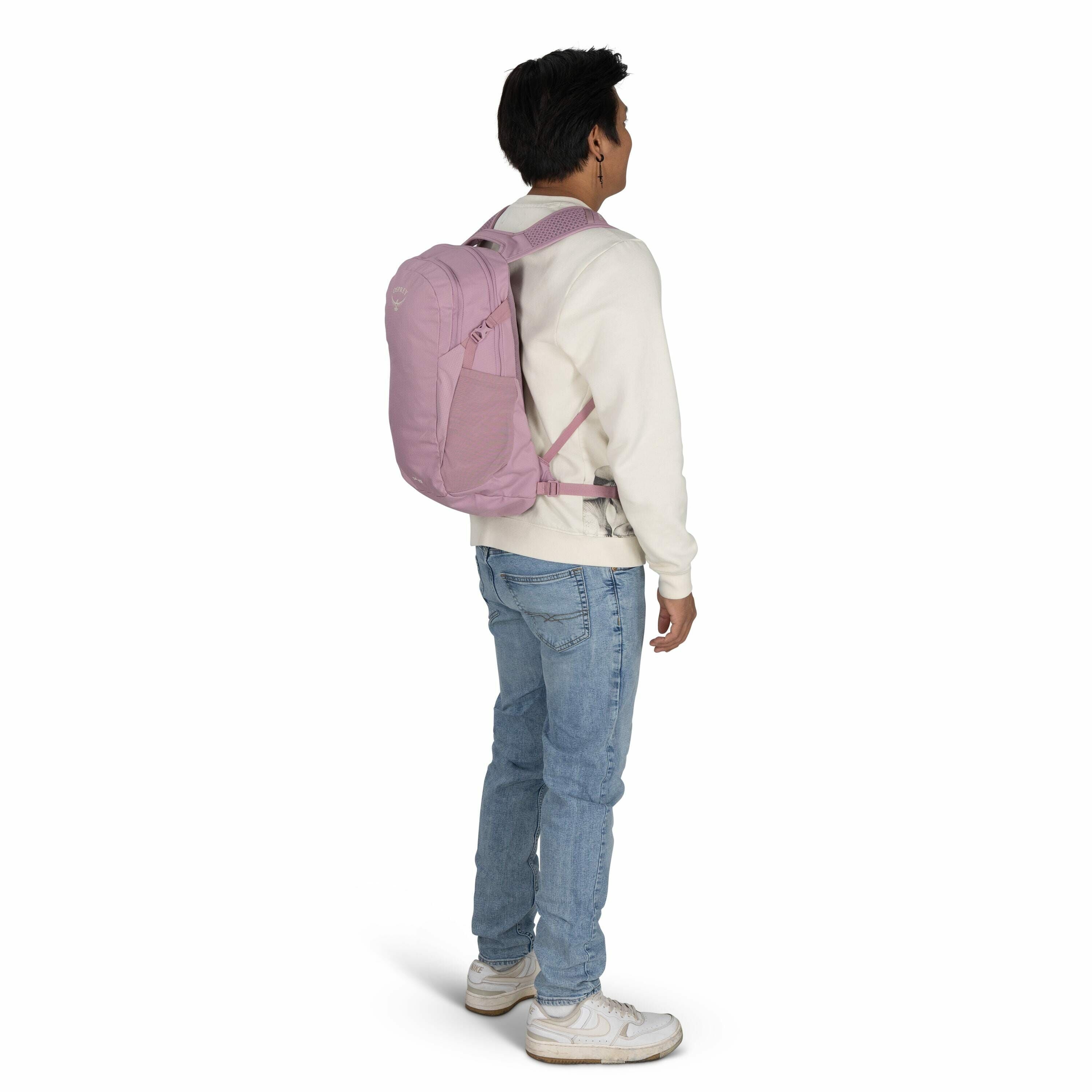 The image depicts a young person standing sideways, modeling a pink backpack. The individual is wearing a white sweater, light blue jeans, and white sneakers, suggesting a casual and relaxed style. The backpack appears to be securely fastened with a chest
