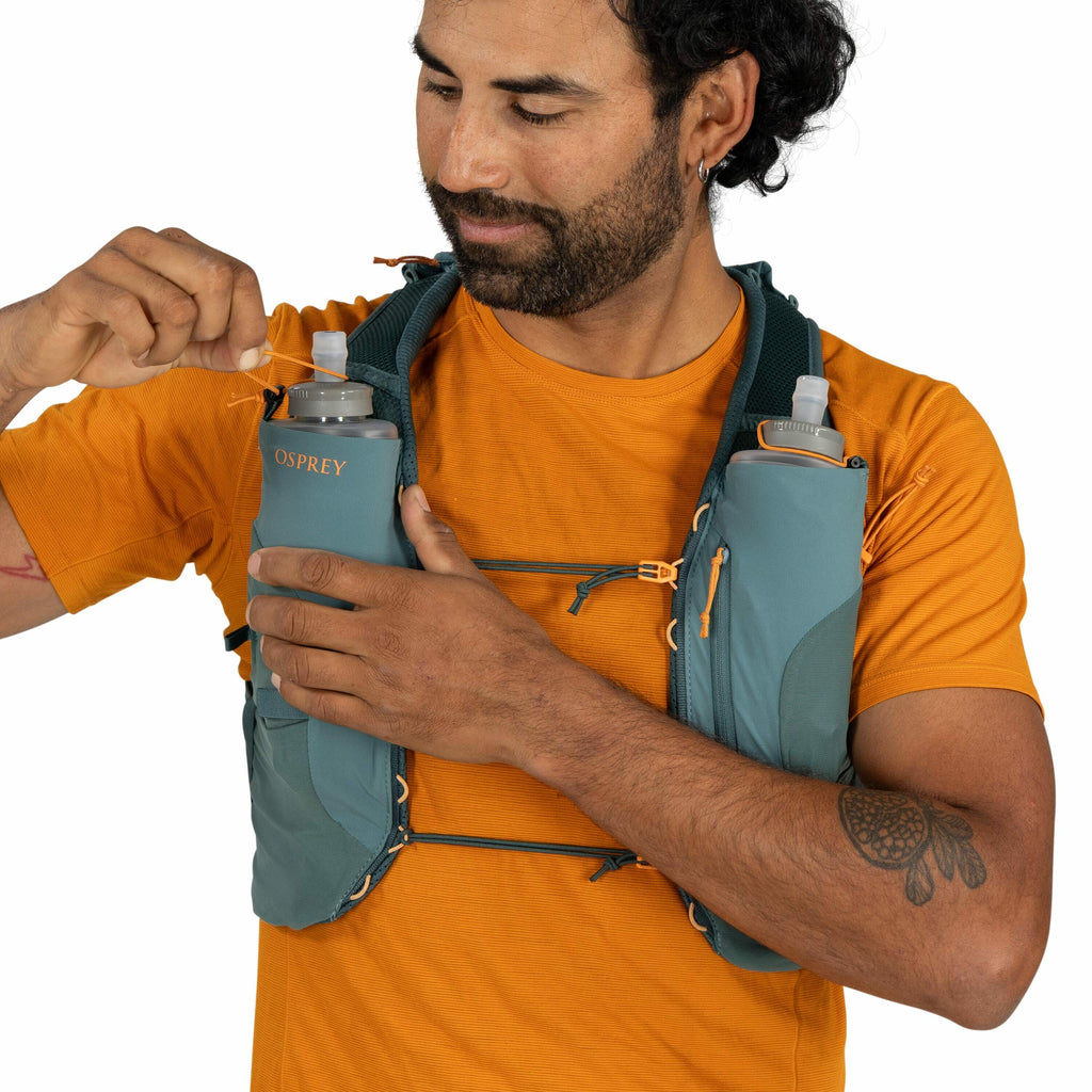 The image shows a person wearing an orange t-shirt and an Osprey running vest. The vest is teal with several compartments, two of which hold grey soft water bottles with spouts. The person is adjusting a strap on the vest, demonstrating its functionality.