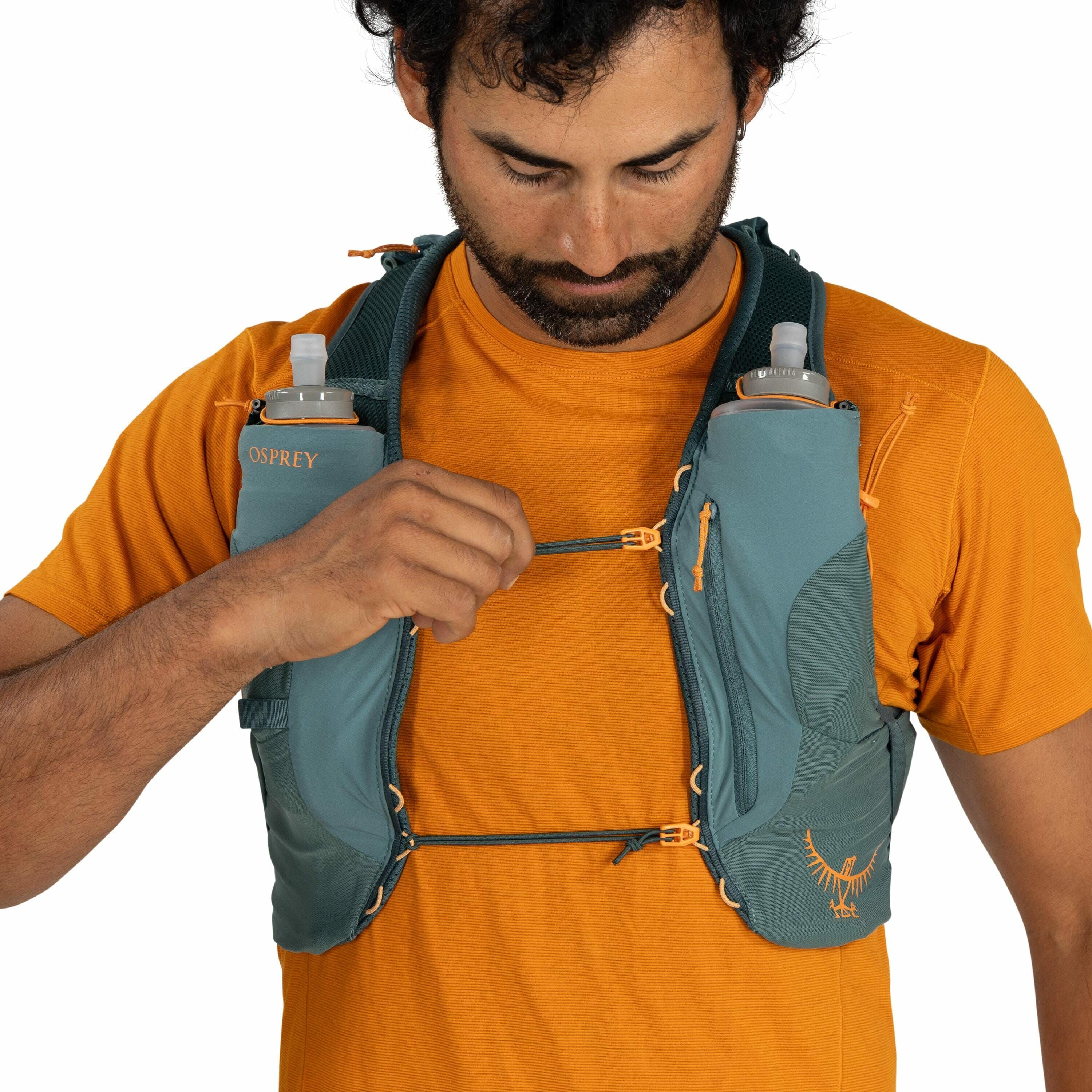 The image shows a person wearing an orange short-sleeved shirt and adjusting the straps of a teal-colored hydration vest. The vest, branded "Osprey," has two front pockets, each holding a soft flask for easy access to water during activities like running