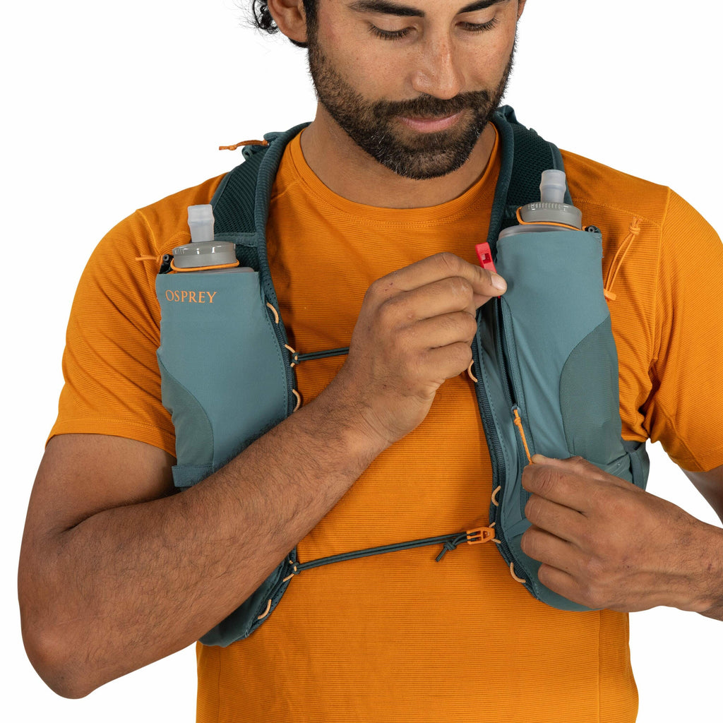 The image shows a person wearing an Osprey running or hydration vest. The vest is teal in color and features two pockets on the chest, each holding a clear water bottle with a nozzle for easy drinking. The person is wearing a bright orange T-shirt underne