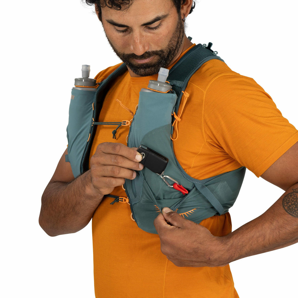The image shows a person wearing an orange shirt and a teal hydration pack vest. The vest features two water bottles in front, secured in bottle pockets. The individual is holding a small black object, attached with a red clip, which could be a compact ca