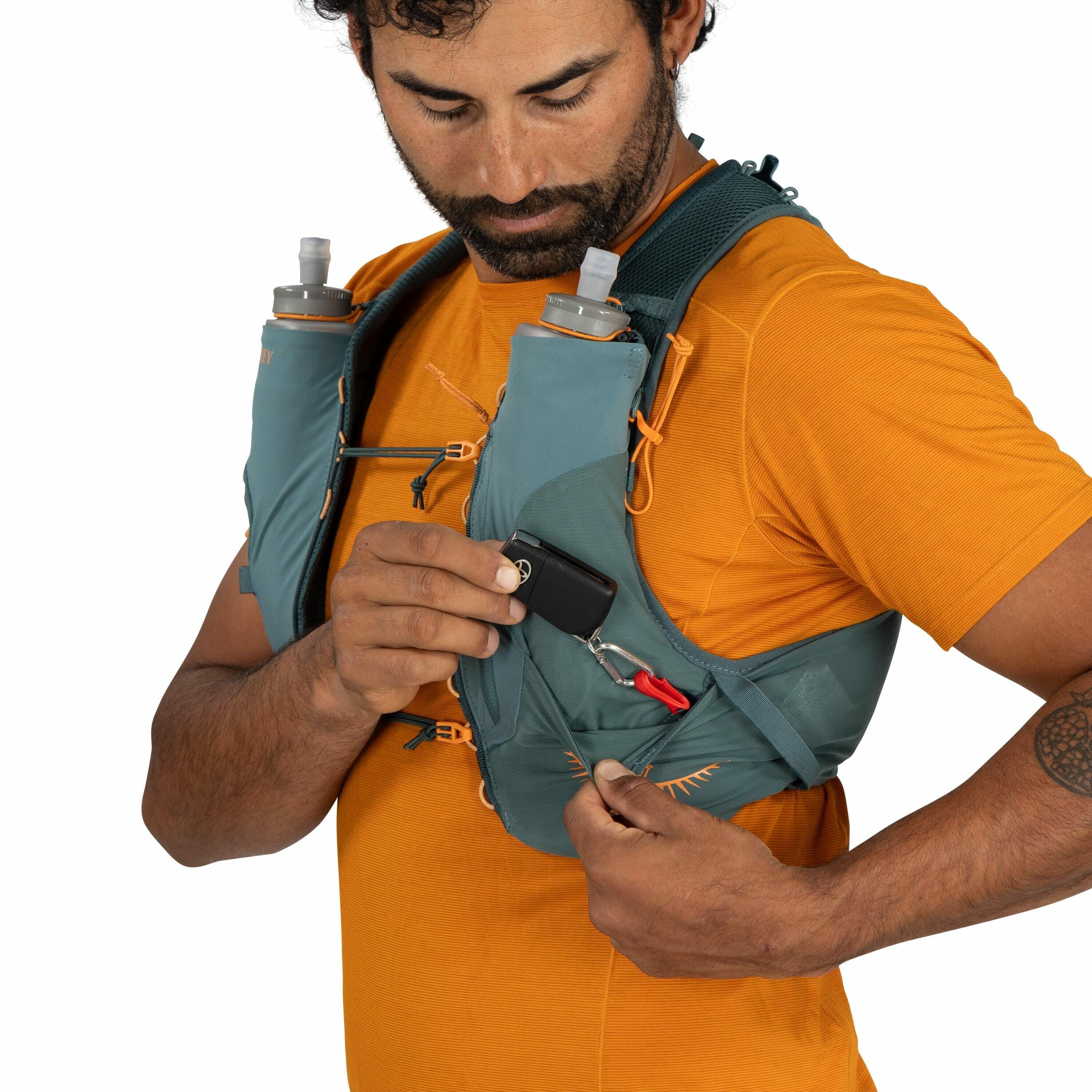 The image shows a person wearing an orange shirt and a teal hydration pack vest. The vest features two water bottles in front, secured in bottle pockets. The individual is holding a small black object, attached with a red clip, which could be a compact ca