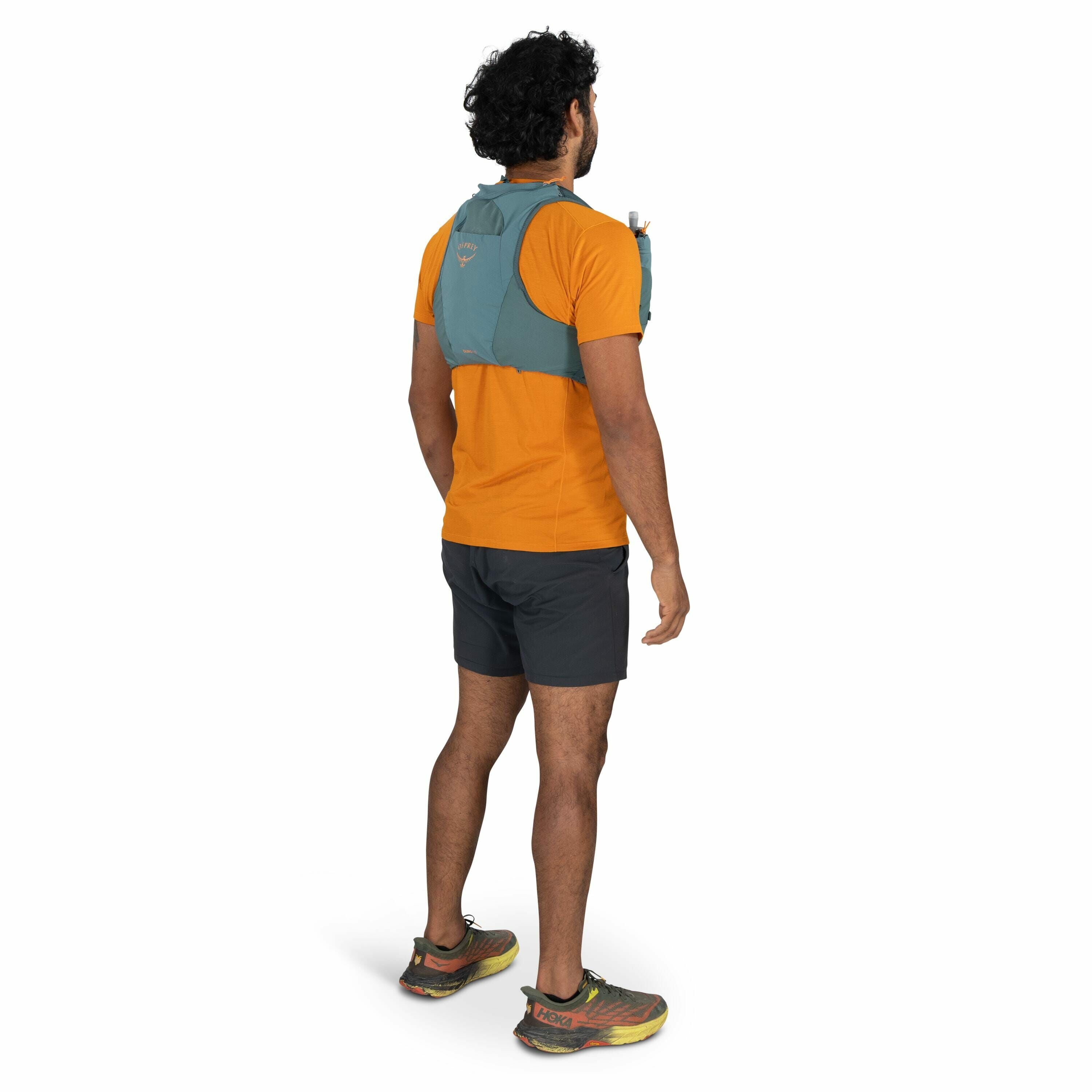 The image features a man standing with his back to the camera, wearing athletic gear. He is dressed in an orange T-shirt and black athletic shorts. On his back, he has a teal hydration vest with a visible straw, indicating readiness for running or hiking
