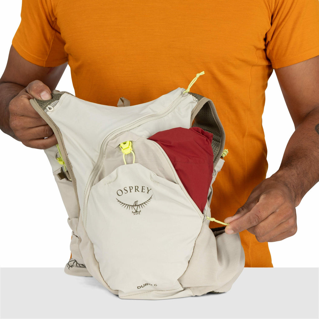 The image shows a person wearing an orange shirt and holding an Osprey backpack. The backpack is beige and has a visible logo that includes the word "Osprey" and a graphic of an osprey bird with outstretched wings. The person is pulling the zipper on a po