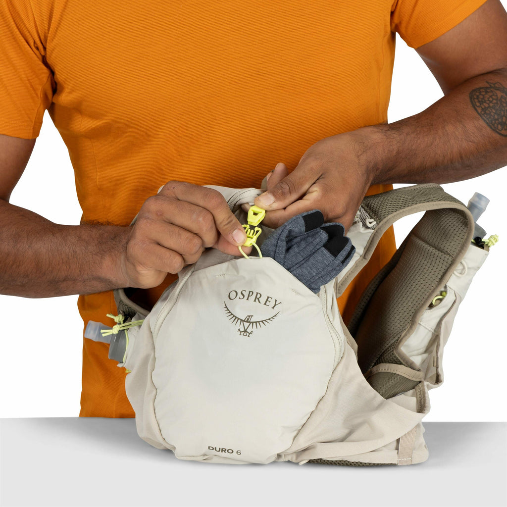The image shows a person wearing an orange shirt, securing the top of an Osprey backpack using a yellow buckle. The backpack, identified as the Osprey Duro 6, is light-colored and has a small logo and branding visible on its front pocket. Inside the pocke