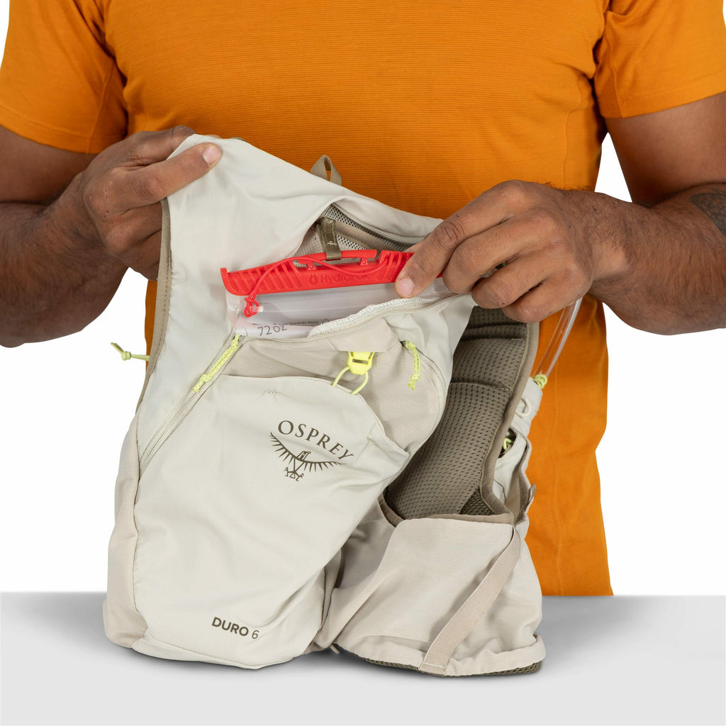 The image depicts a person wearing an orange shirt, holding a light beige Osprey Duro 6 backpack in their hands. The backpack is partially opened, revealing a hydration reservoir with a red top marked with "Hydrapak". The person is holding the hydration r