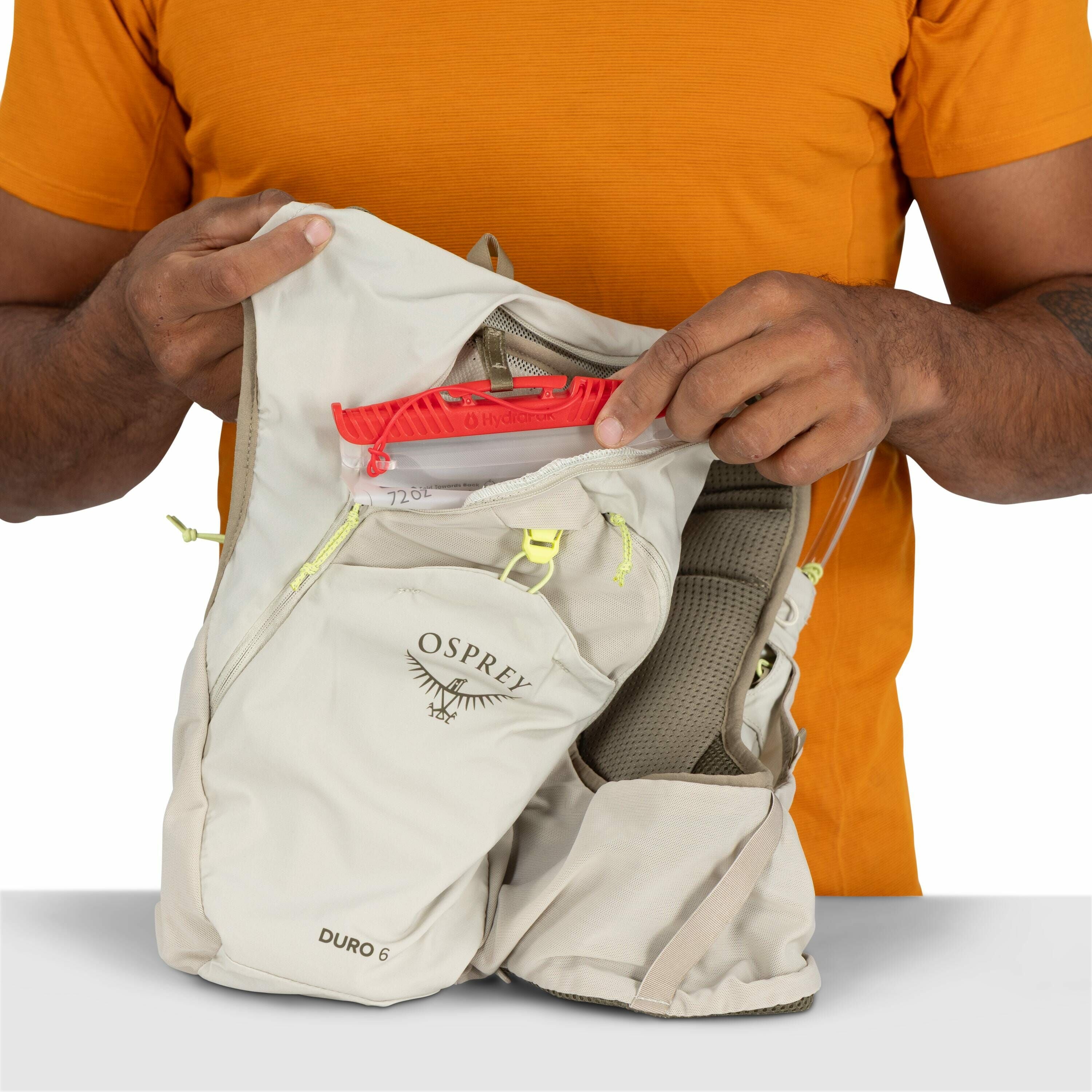The image depicts a person wearing an orange shirt, holding a light beige Osprey Duro 6 backpack in their hands. The backpack is partially opened, revealing a hydration reservoir with a red top marked with "Hydrapak". The person is holding the hydration r