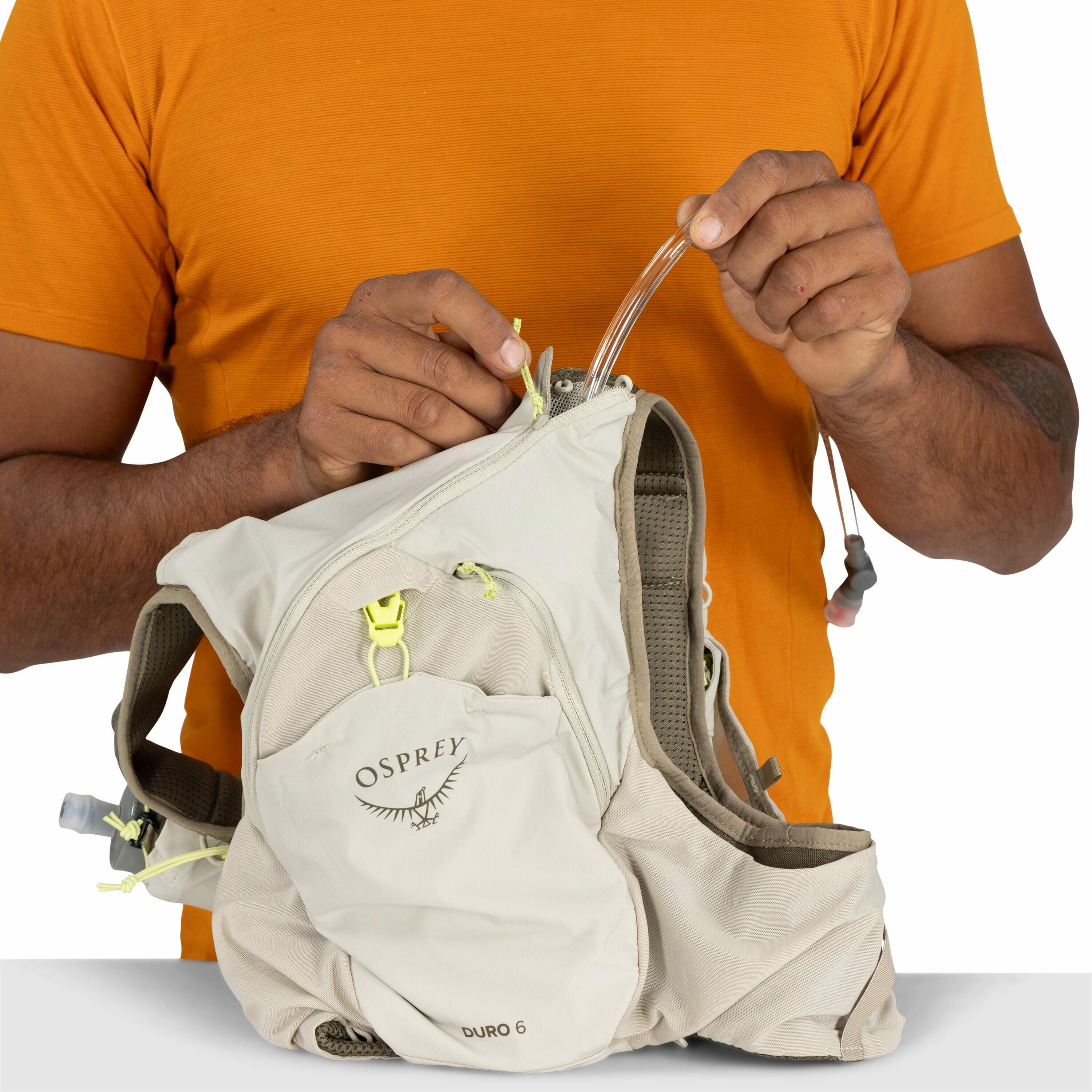 The image shows a person holding an Osprey Duro 6 hydration vest. The vest is primarily light beige with the Osprey logo and "DURO 6" visible on one of the front pockets. The person, wearing an orange shirt, is seen adjusting or managing the hydration tub