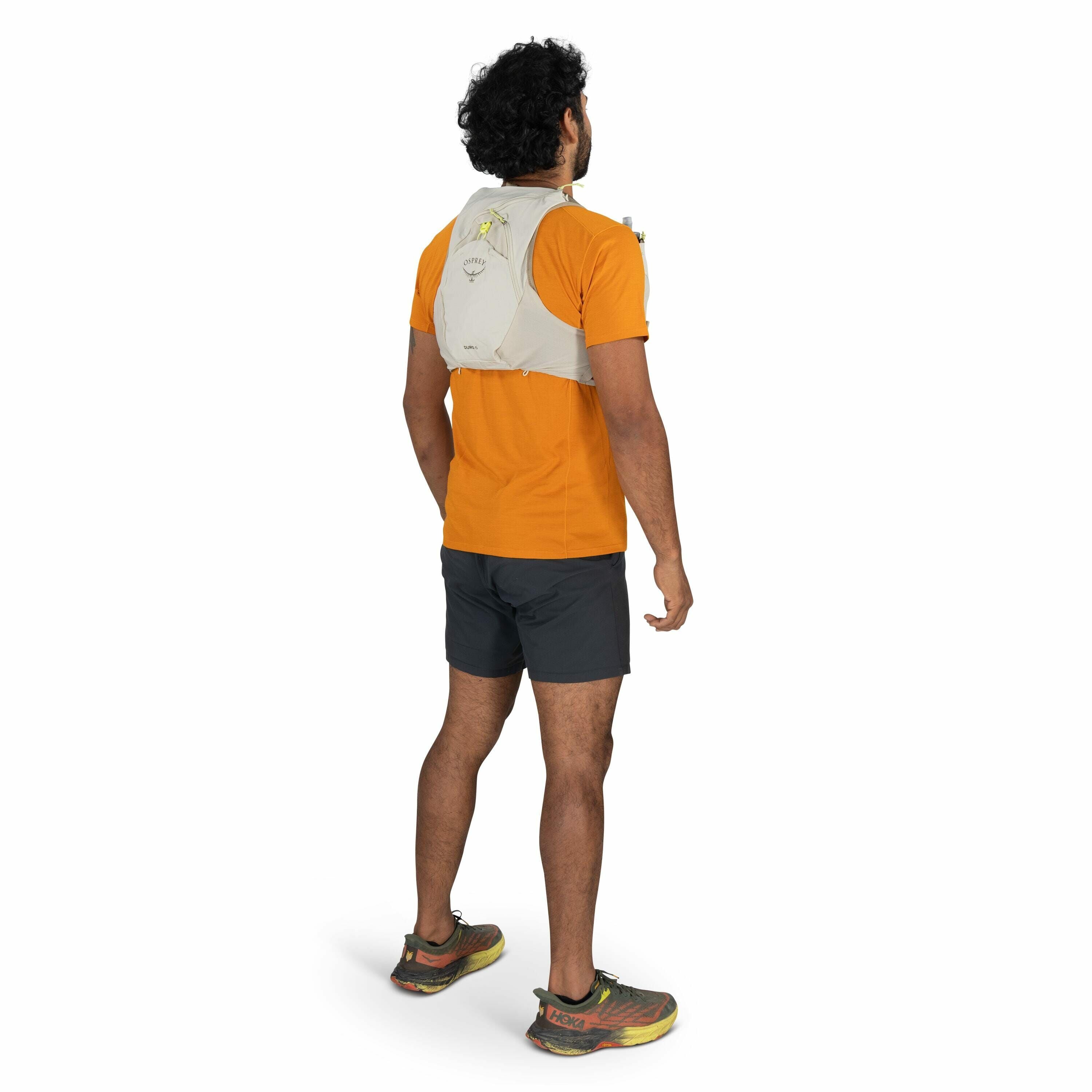 The image shows a man standing and facing away from the camera, showcasing a beige running vest. The vest appears to be designed for long-distance running or trail running, and it has multiple pockets for storage, as seen by items protruding from them. Th