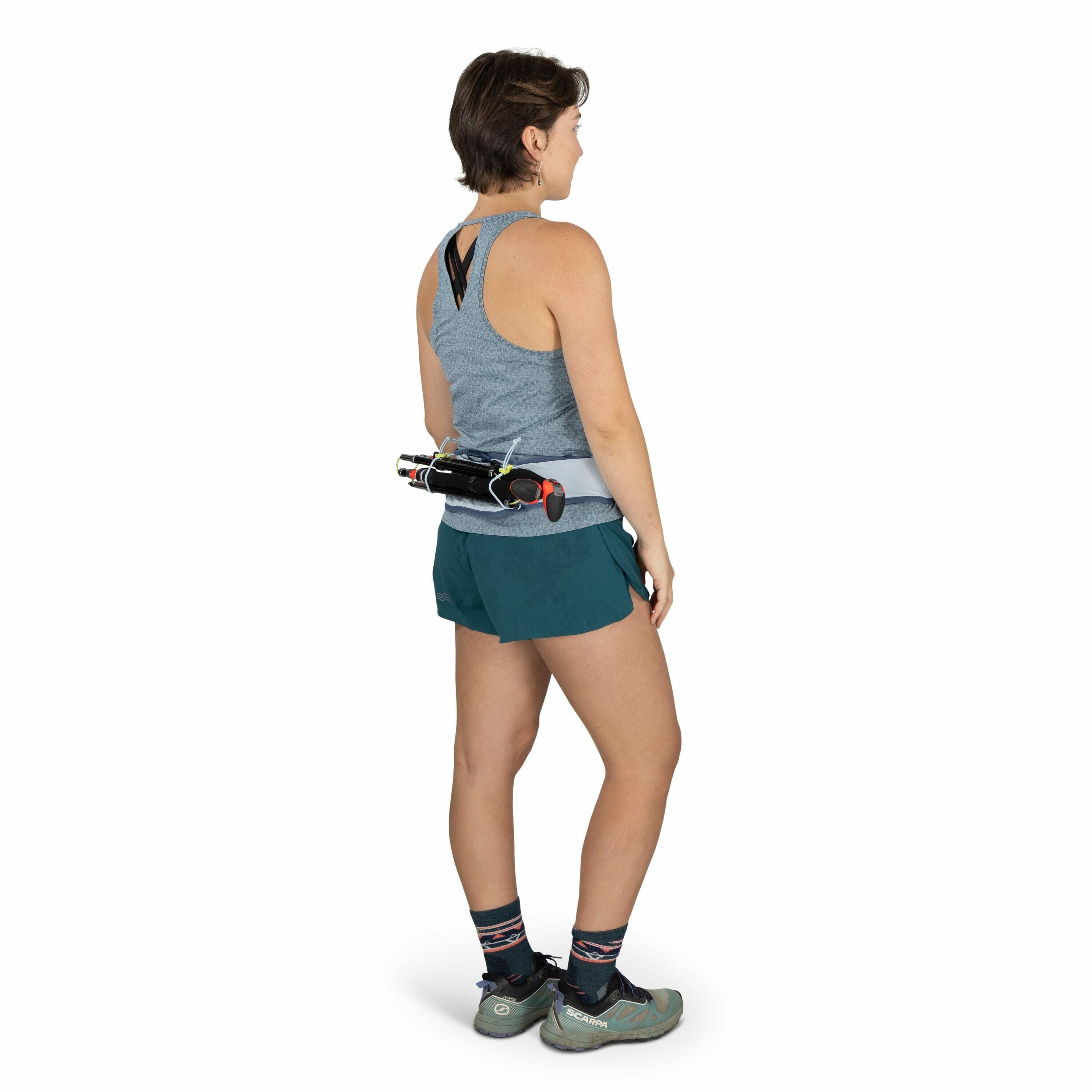 The image features a person standing in a side pose, wearing athletic attire suitable for running or hiking. The person is dressed in a gray sleeveless top with a cutout detail at the back, paired with teal shorts. On their feet are a pair of light green