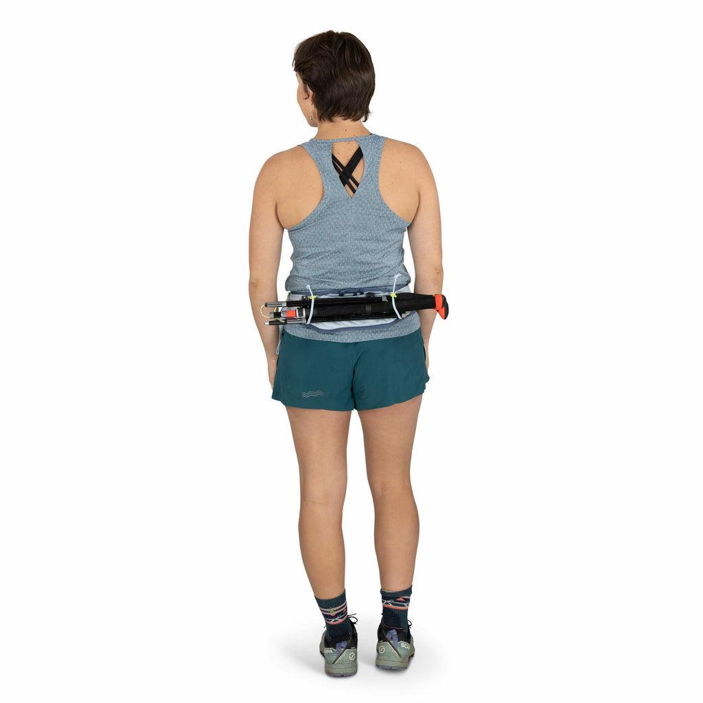 The image shows a woman standing and facing away from the camera, dressed in athletic attire. She is wearing a light blue sleeveless top with a crisscross design at the back and teal shorts with a subtle logo on the side. On her feet are multicolored sock