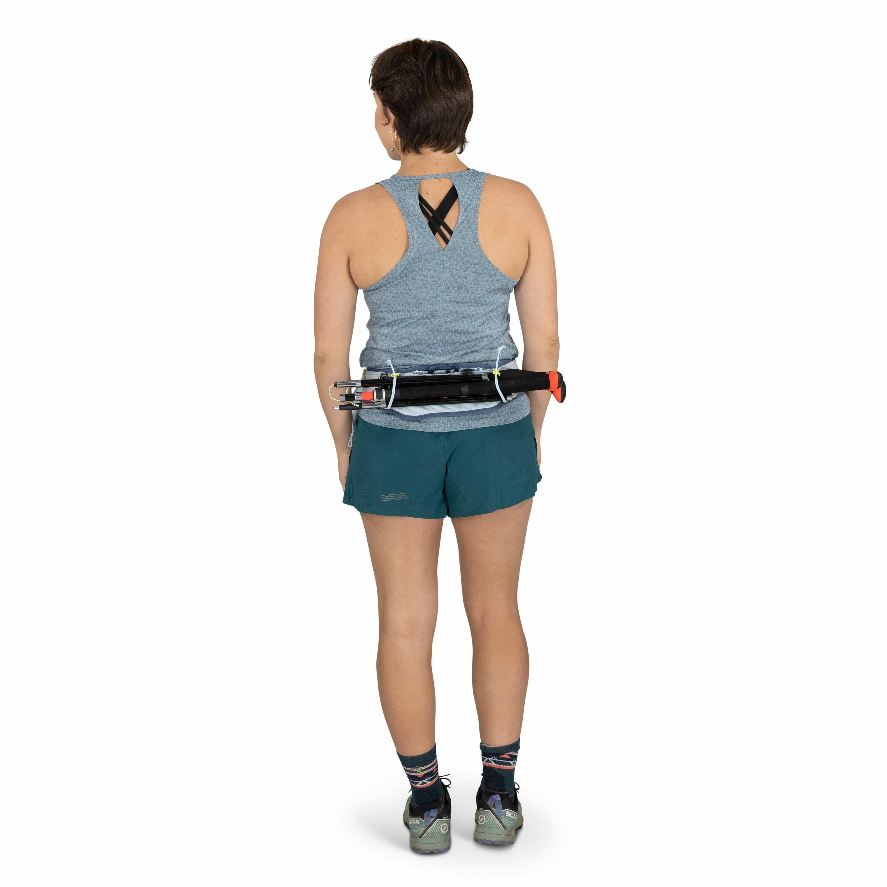 The image shows a woman standing and facing away from the camera, dressed in athletic attire. She is wearing a light blue sleeveless top with a crisscross design at the back and teal shorts with a subtle logo on the side. On her feet are multicolored sock