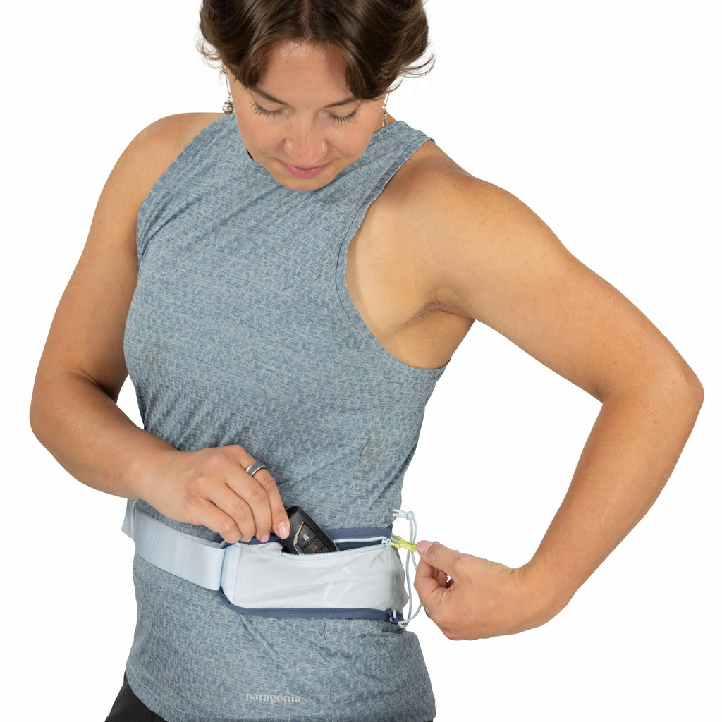 The image shows a woman wearing a light blue textured sleeveless top and a waist bag. The bag is secured around her waist and has a pouch where she is placing a walkie-talkie-like device. The bag is light gray with a hint of blue, and it has a bright yell