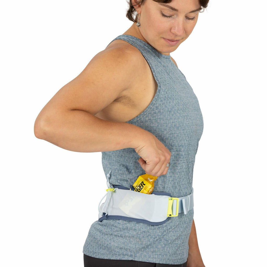 The image shows a person wearing a blue sleeveless top and dark pants, standing in profile with their right arm bent. The person has a small blue and white waist belt with a mesh pouch that is fastened around their waist. The pouch has a bright yellow zip