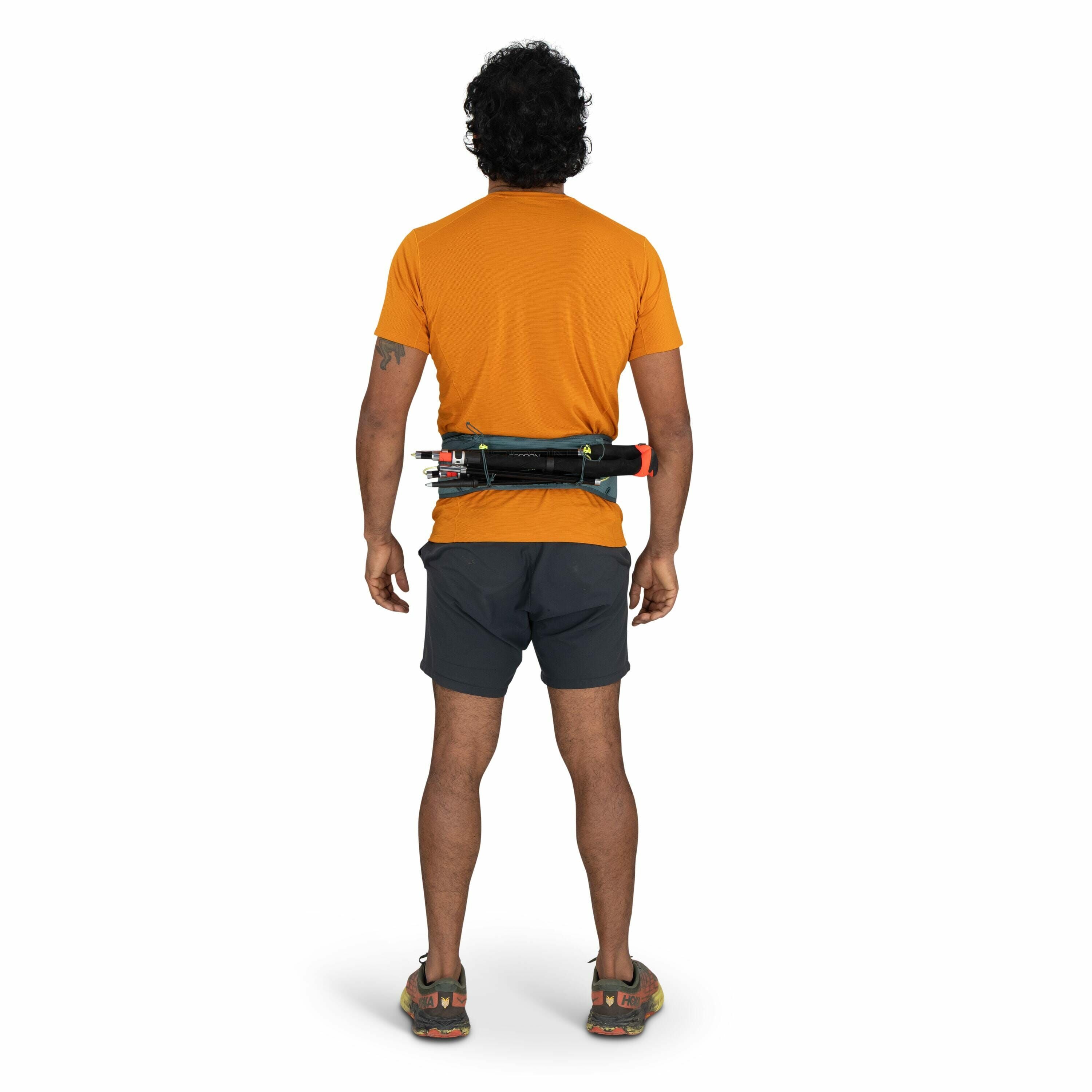 The image features a man standing with his back to the camera. He is wearing an orange t-shirt, dark grey shorts, and sports shoes. Around his waist, he has a black belt or waist pack that appears to be equipped with various items, possibly tools or gear