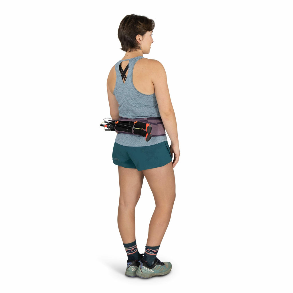 The image depicts a person standing, facing away from the camera, wearing athletic gear suitable for outdoor activities. They are dressed in a light gray, sleeveless athletic top with a cross-strap design at the back and teal shorts. The individual is wea
